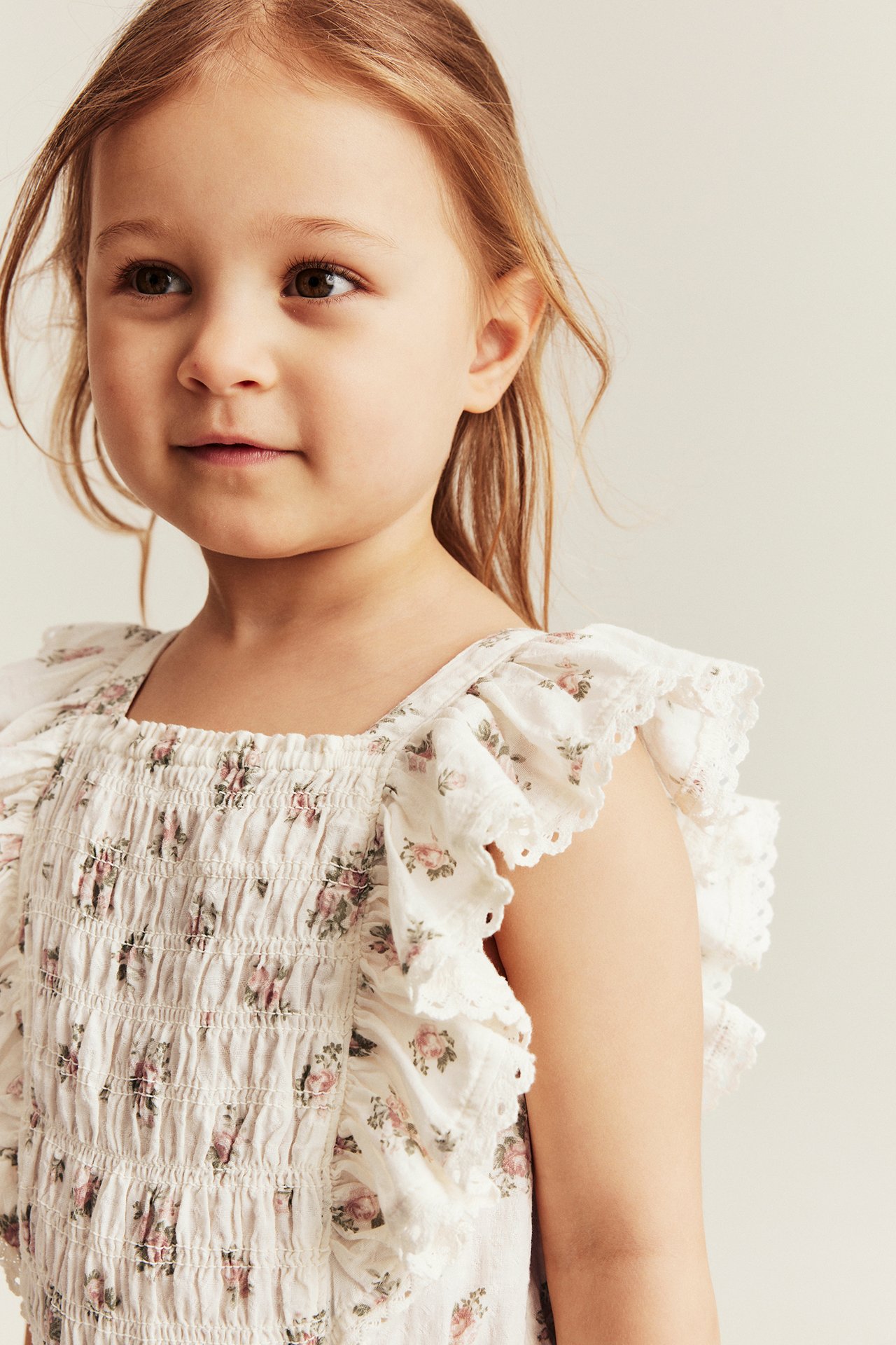 Close-up of a kid wearing an off-white floral jumpsuit with smocking and ruffled, lace-trimmed sleeves.