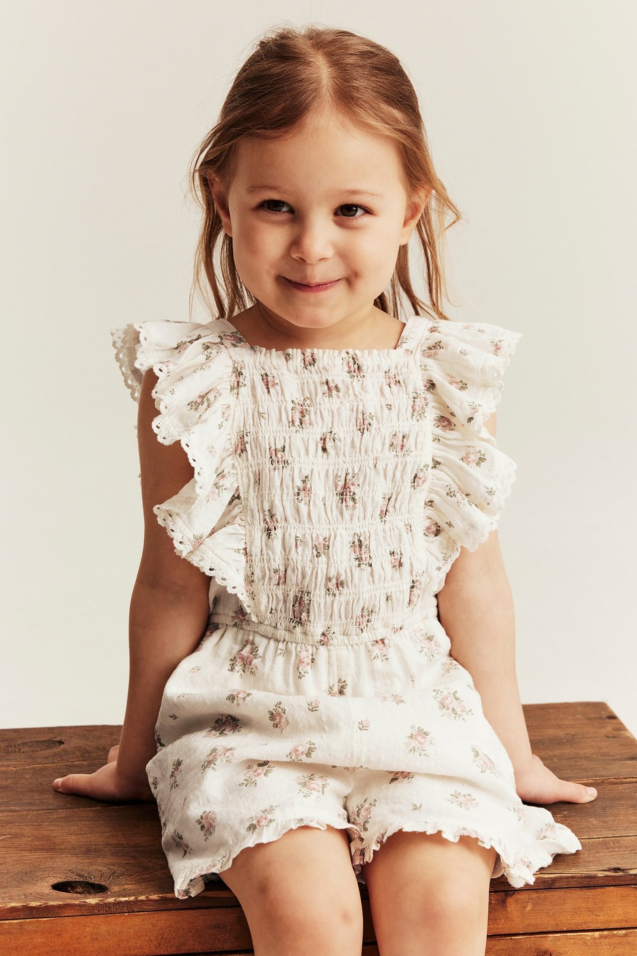 Front view: Kid wears offwhite floral short-legged jumpsuit with smocked bodice, ruffled lace shoulders.