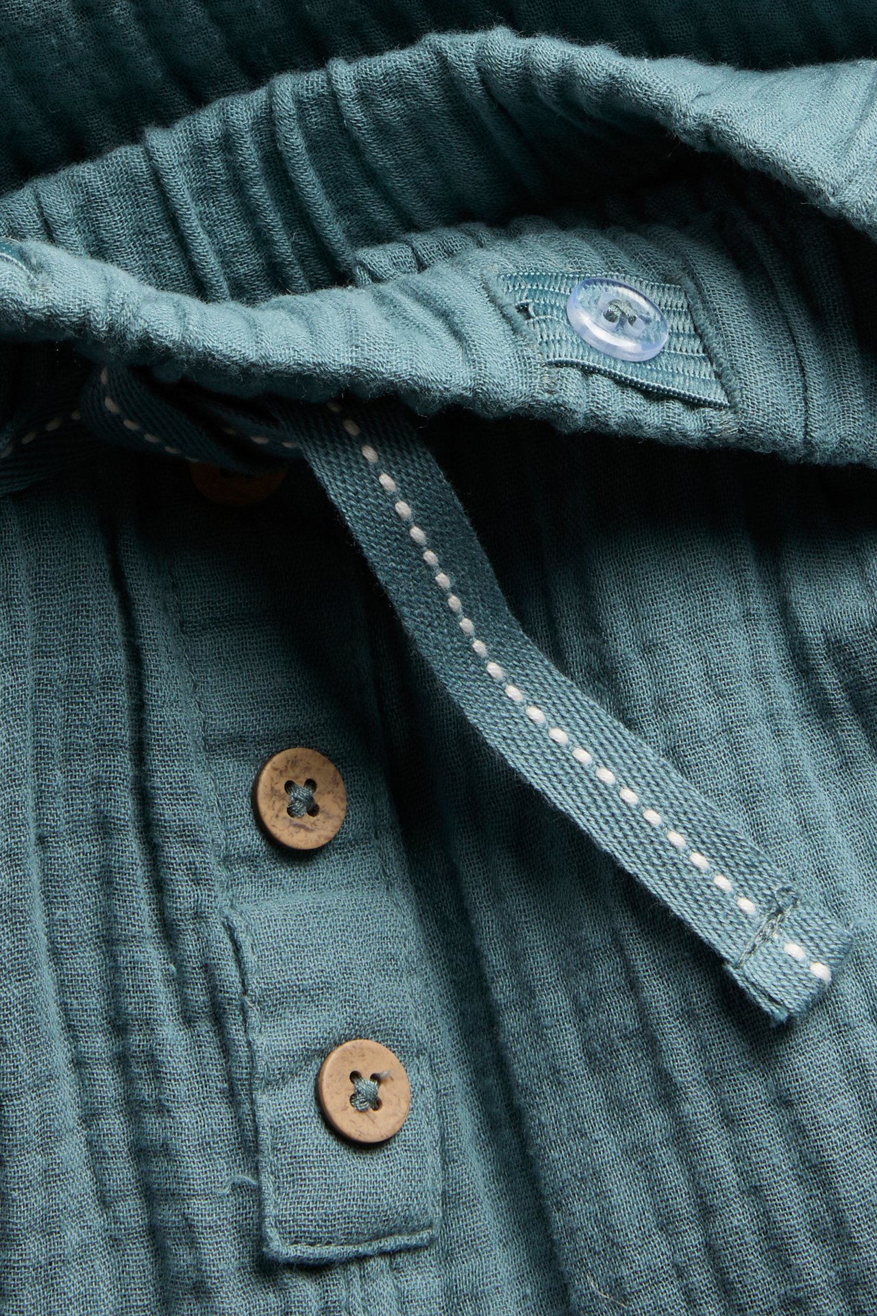 Nahaufnahme von dunkel türkisen Baumwoll-Musselin-Hosen mit Gummizugbund, Fischgrät-Kordelzug und dekorativen Holzknöpfen.