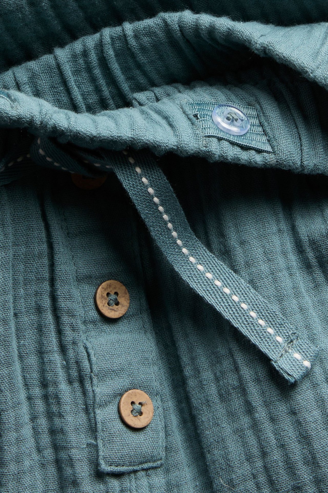 Nahaufnahme von dunkel türkisen Baumwoll-Musselin-Hosen mit Gummizugbund, Fischgrät-Kordelzug und dekorativen Holzknöpfen.