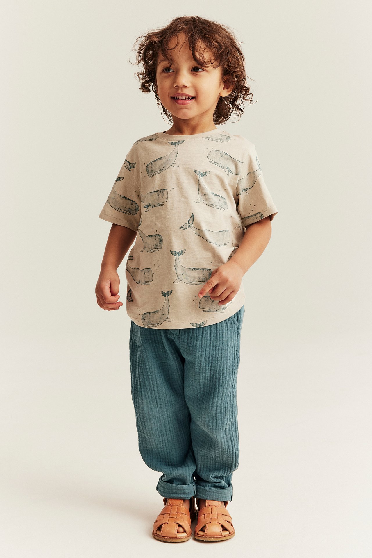 Vorderansicht: Kind trägt dunkel-türkise Musselin-Hose, beiges T-Shirt mit Walmuster und braune Sandalen.