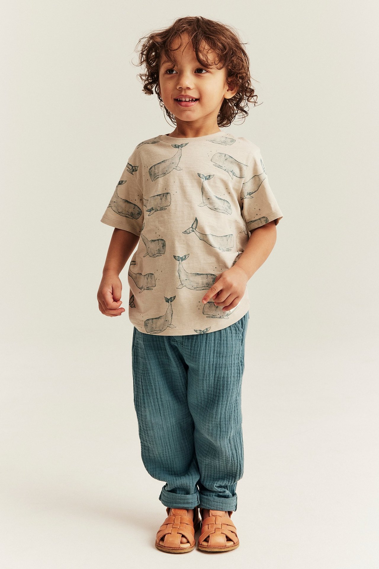 Vorderansicht: Kind trägt dunkel-türkise Musselin-Hose, beiges T-Shirt mit Walmuster und braune Sandalen.