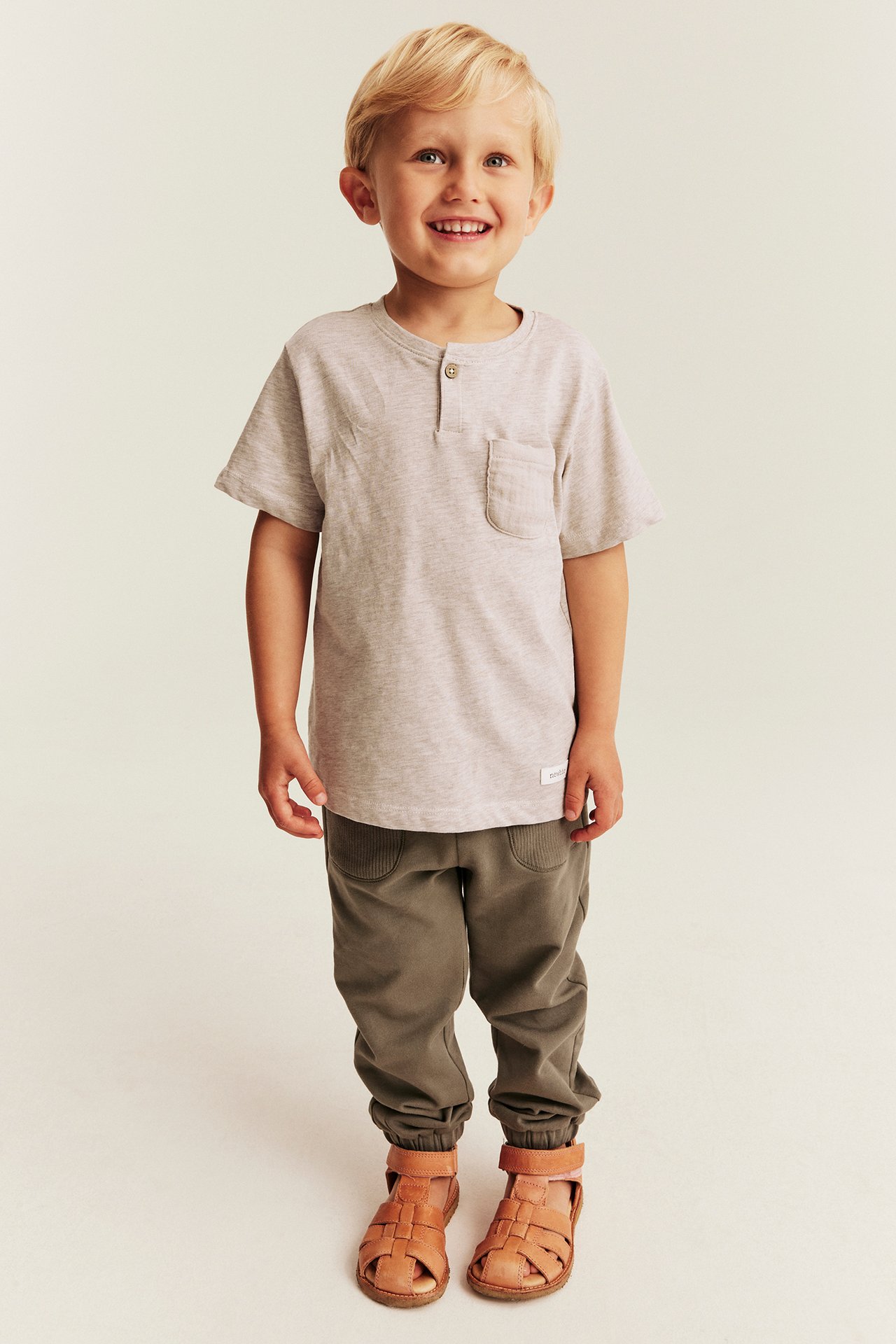 Vorderansicht eines Kindes in hellgrauem Henley-T-Shirt, dunkeloliven Jogger und braunen Ledersandalen.