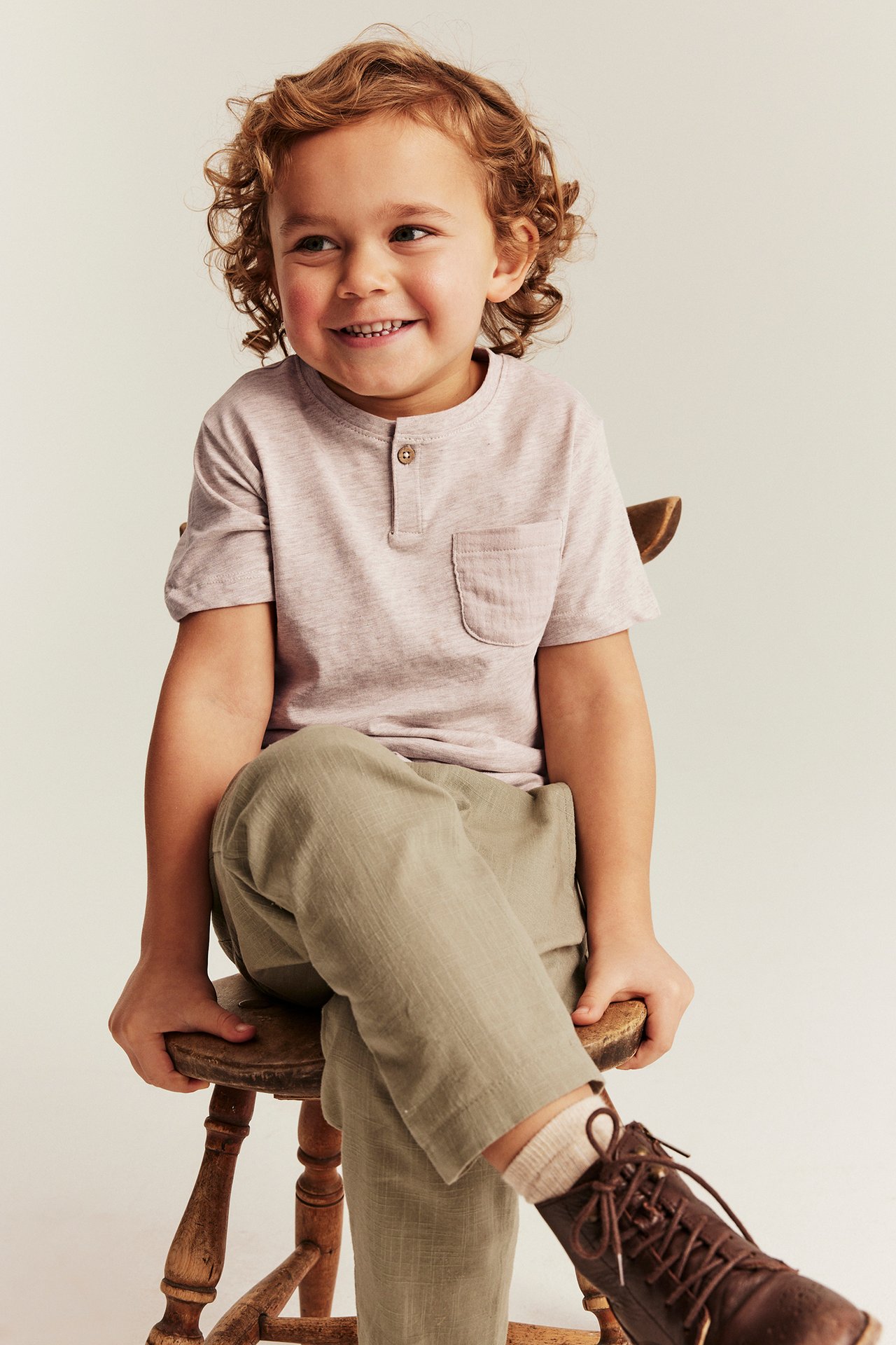Vorderansicht: Kind trägt ein beiges, meliertes T-Shirt mit Knopfleiste, Brusttasche, olivgrüne Hose, braune Stiefel.
