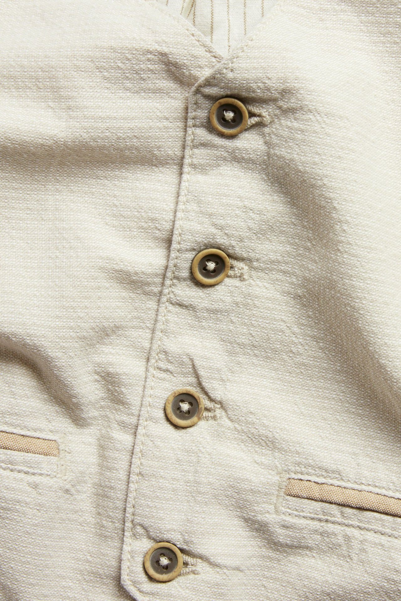 Nahaufnahme: Beige gewebte Baumwollweste mit braunen Knöpfen und Ziertaschen; gestreiftes Futter sichtbar.