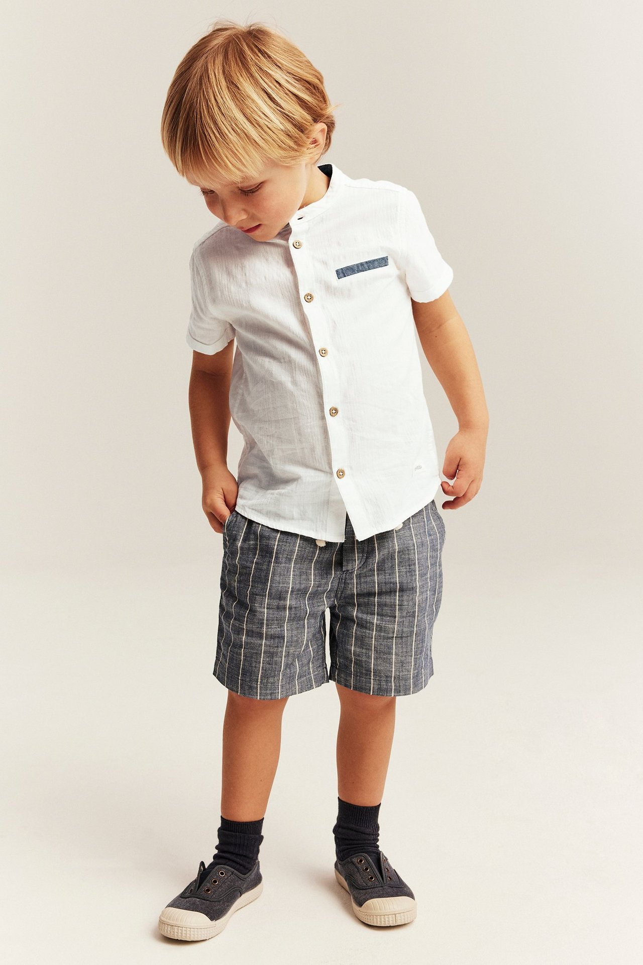 Vorderansicht eines Kindes in einem cremeweißen kurzärmeligen Hemd, dunkelblauen karierten Shorts, dunklen Socken und Turnschuhen.