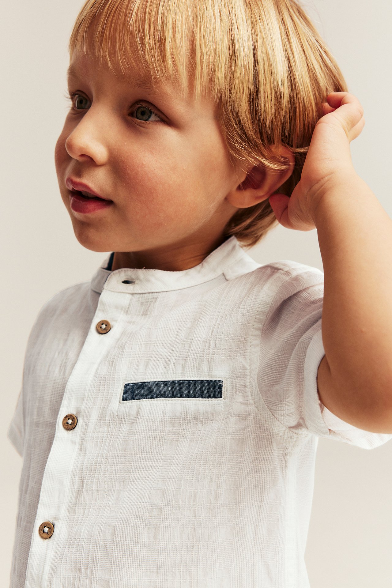 Close-up of a kid in an offwhite short-sleeved woven shirt with band collar, brown buttons, chambray pocket.