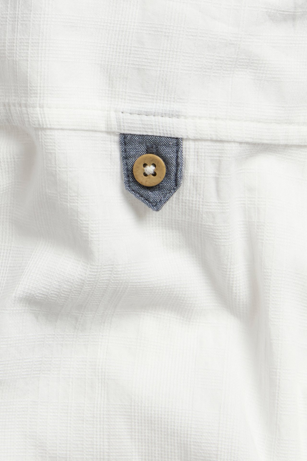 Nahaufnahme eines cremeweißen gewebten Baumwollhemdes mit dezenter Gitterstruktur und einem blauen Chambray-Riegel mit Holzknopf.