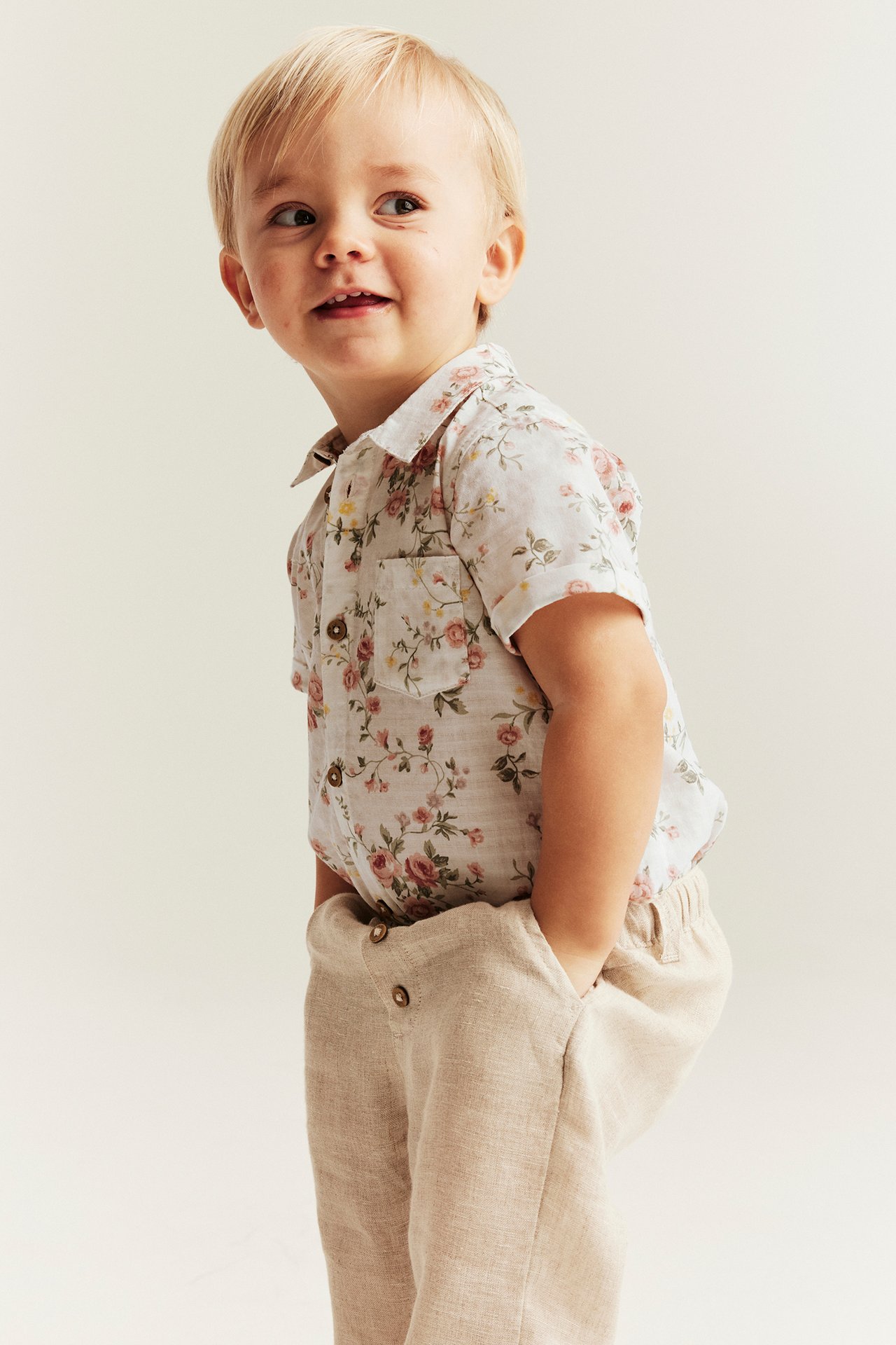 Front-side view of a kid wearing an offwhite pink floral short-sleeved cotton shirt-bodysuit and beige trousers.