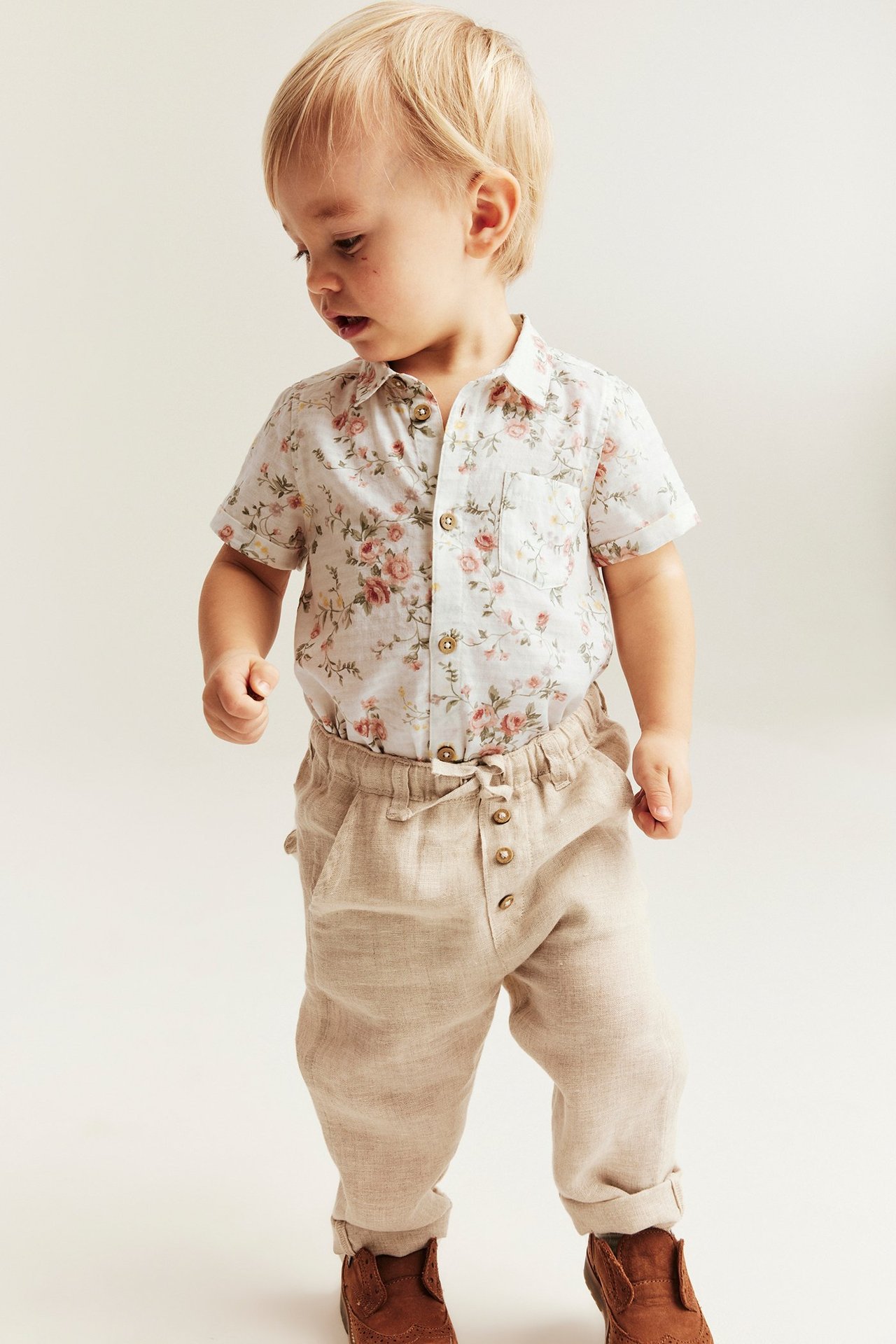 Front view of a baby wearing an off-white floral short-sleeved cotton shirt body and beige trousers.
