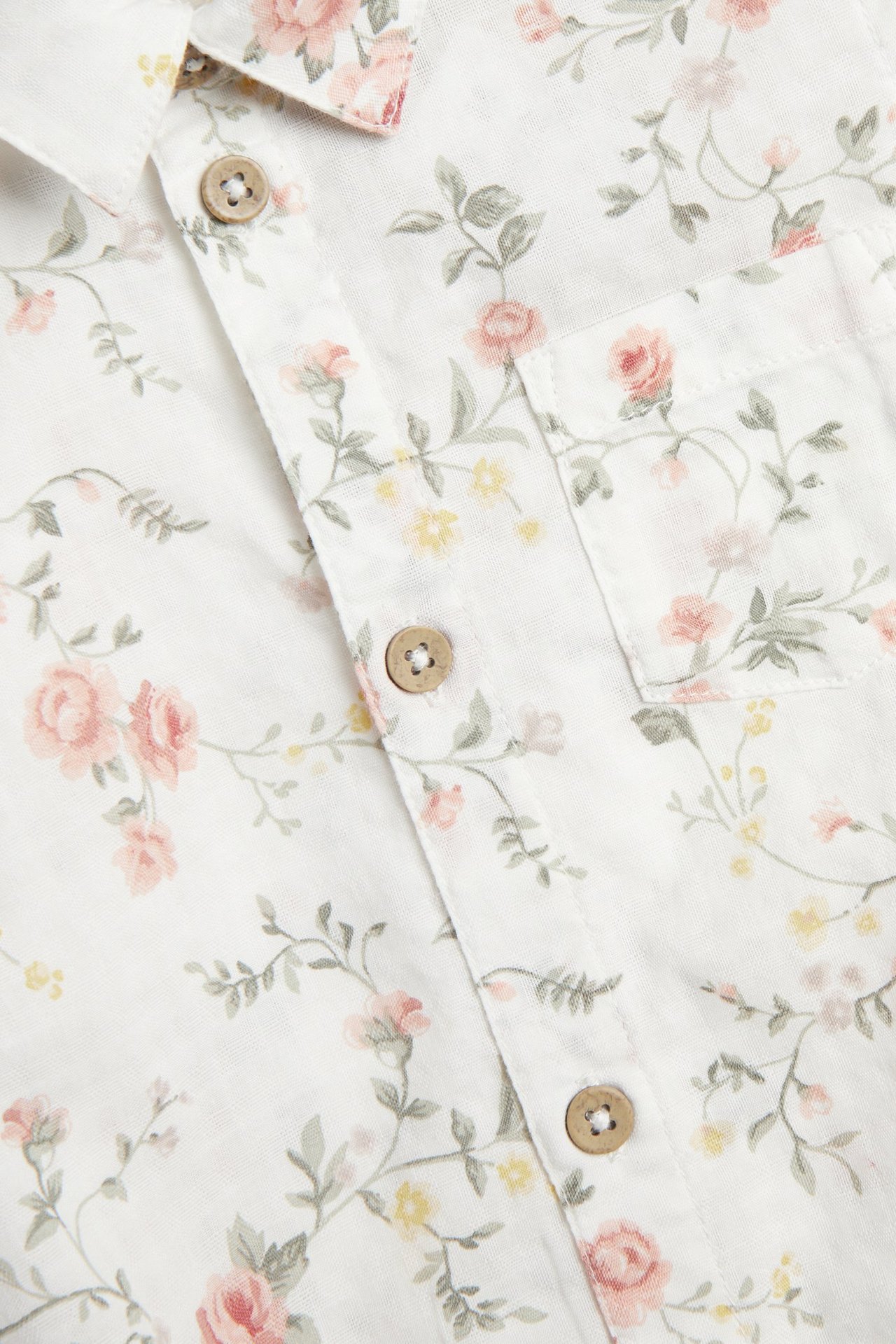 Nahaufnahme eines offwhite geblümten kurzärmligen Baby-Hemdbodys mit Holzknöpfen und Brusttasche.