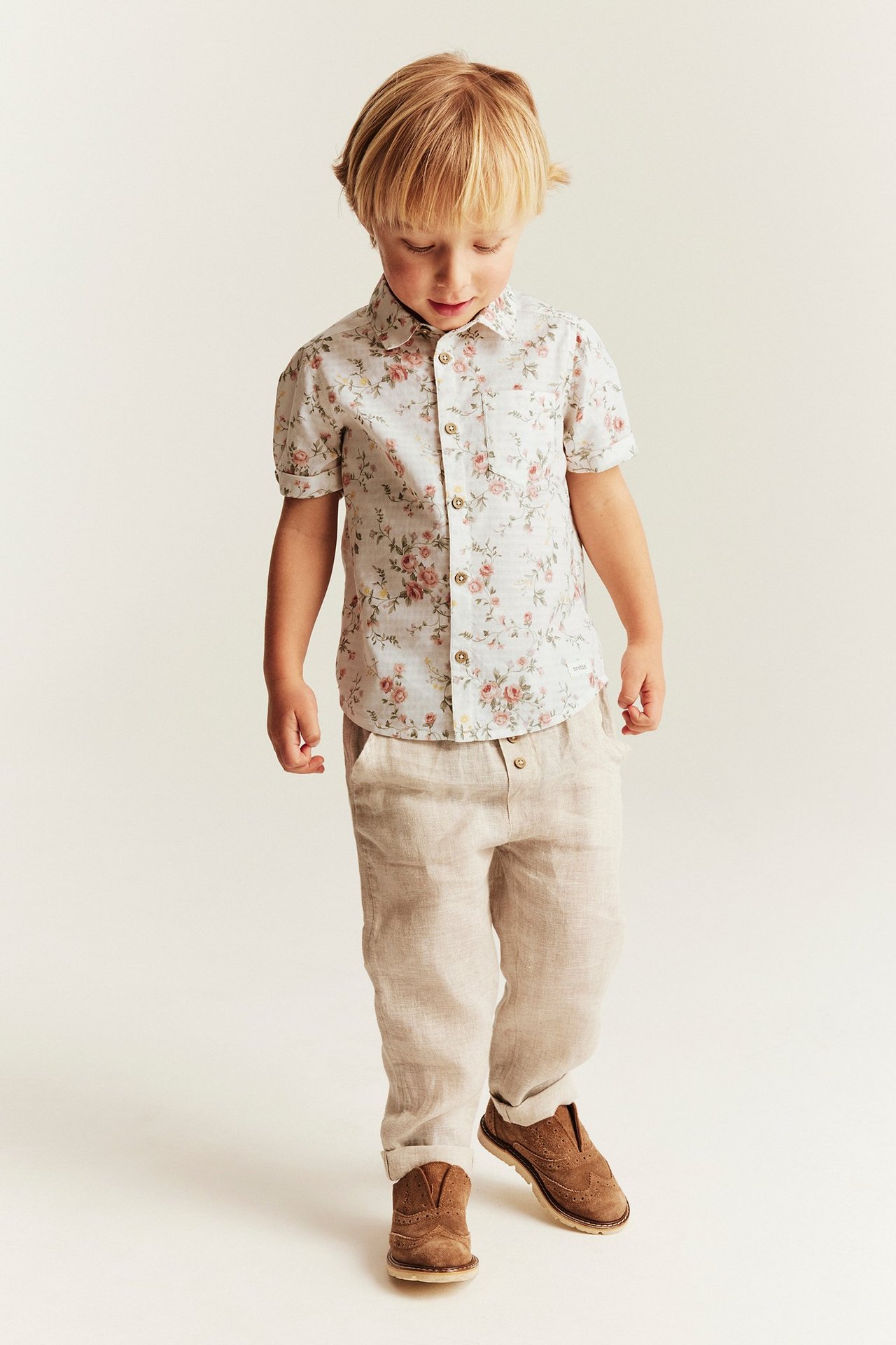 Vorderansicht: Kind trägt offwhite kurzärmeliges Blumen-Baumwollhemd, hellbeige Hose und braune Schuhe.