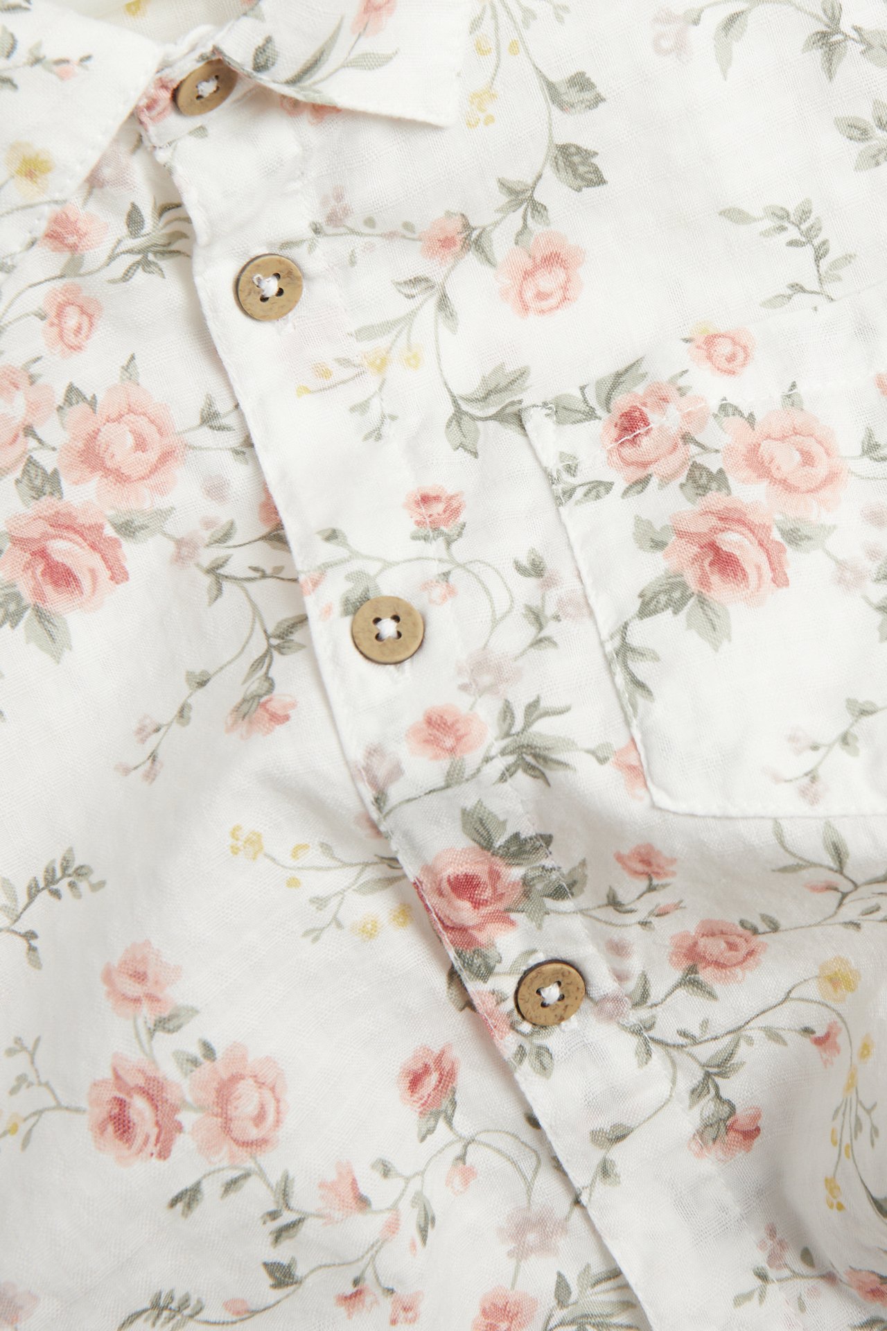 Close-up front: Off-white cotton short-sleeved shirt with pink floral print, collar, wooden buttons, chest pocket.