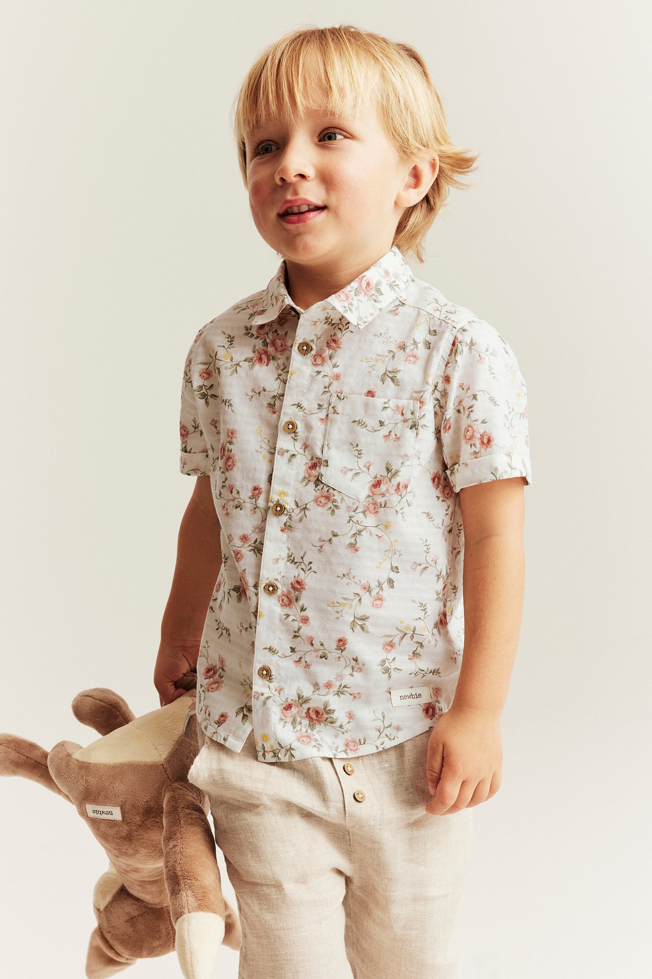 Front view of a kid wearing an offwhite floral cotton shirt, light beige trousers, holding a brown animal.