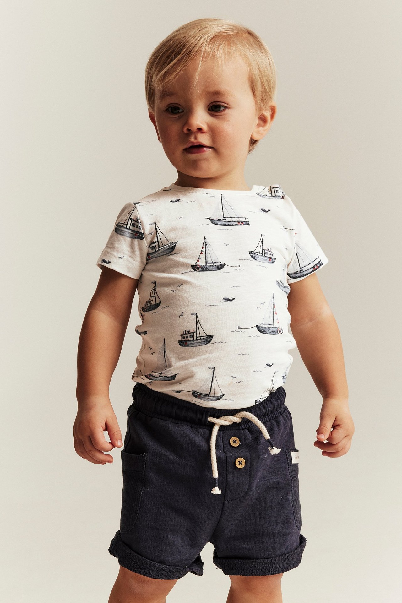 Vorderansicht eines Babys in einem cremefarbenen, kurzärmeligen Baumwoll-Body mit Segelbootmuster und dunkelblauen Shorts.