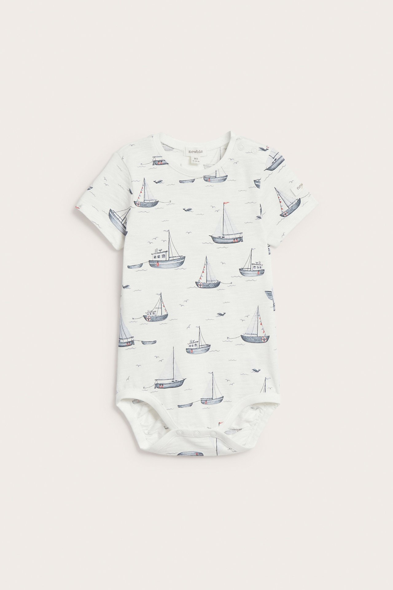 Front view of an off-white baby short-sleeved bodysuit with an all-over sailboat print and snap buttons.