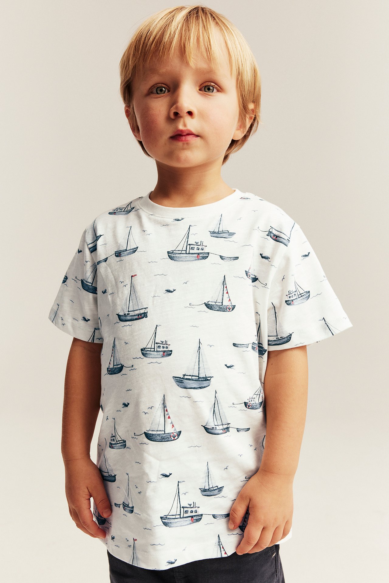 Front view of a kid wearing an off-white short-sleeved cotton t-shirt with a blue sailboat pattern.