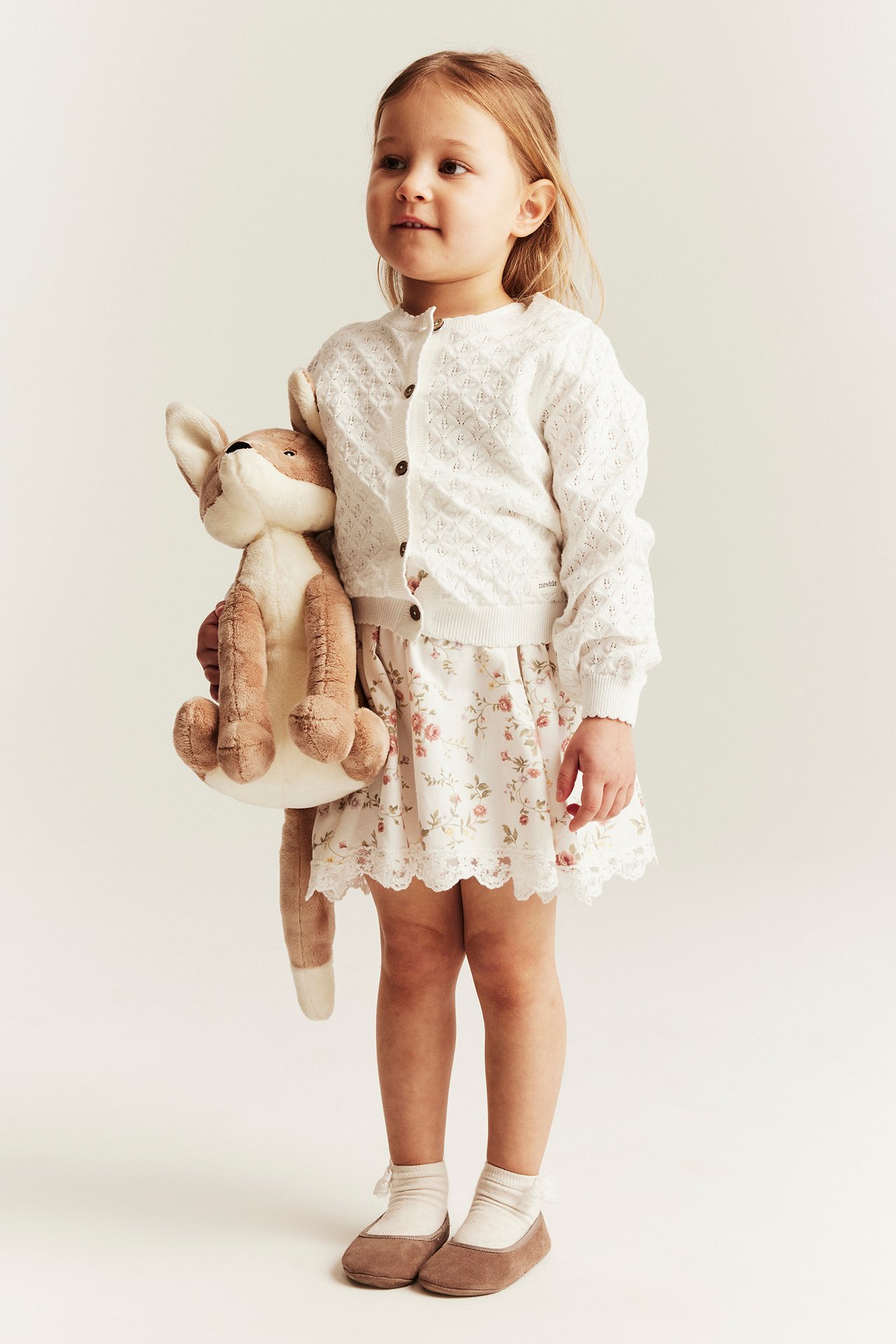 Front view: Kid in an offwhite patterned knit cotton cardigan with buttons, floral skirt, and brown fox toy.