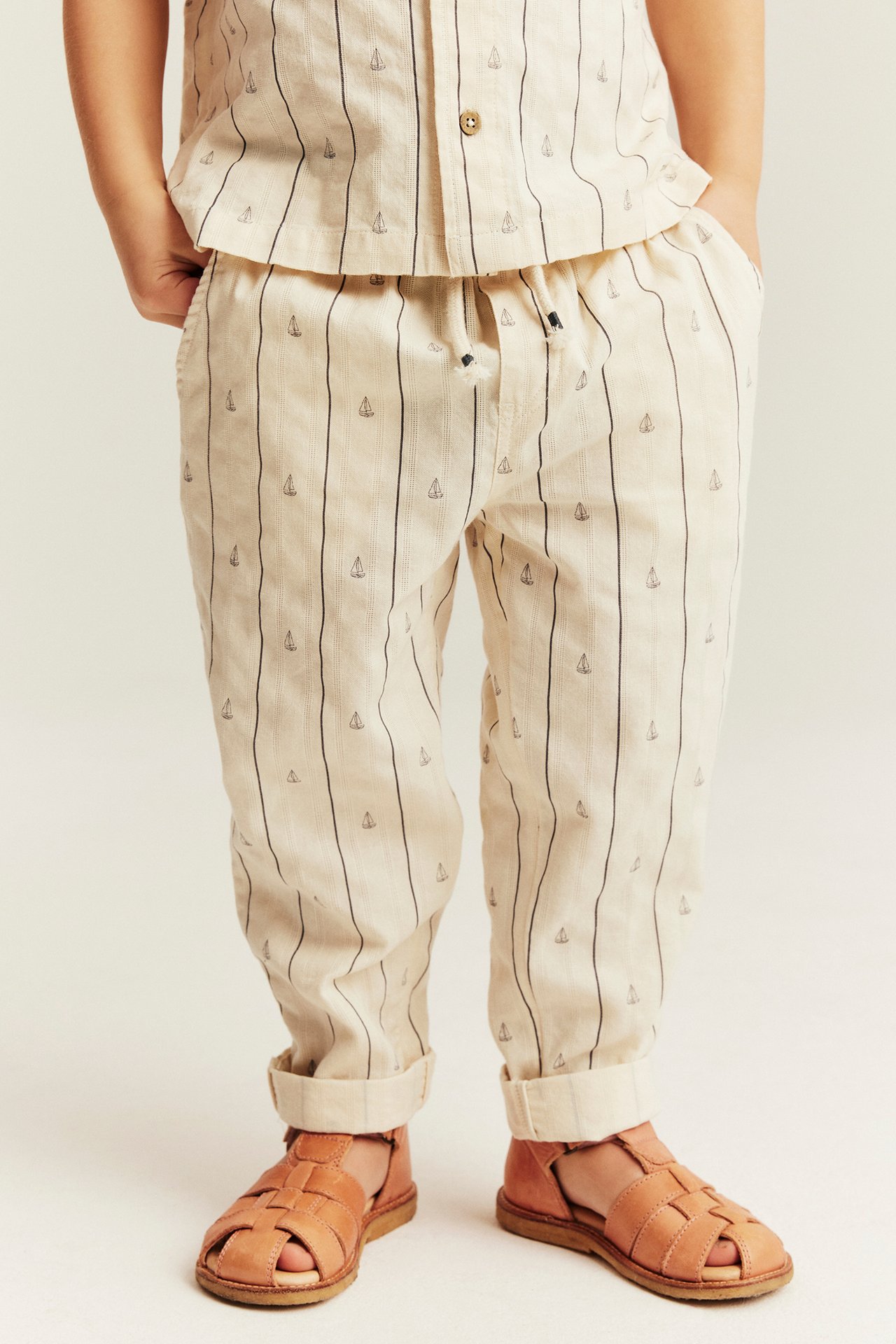 Vorderansicht eines Kindes in cremefarbener gestreifter Hose mit Segelbooten, passendem Hemd und braunen Sandalen.