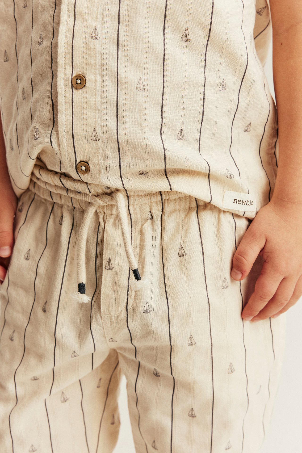 Kid wears patterned white cotton trousers with stripes, sailboats, drawstring waist, and matching shirt.