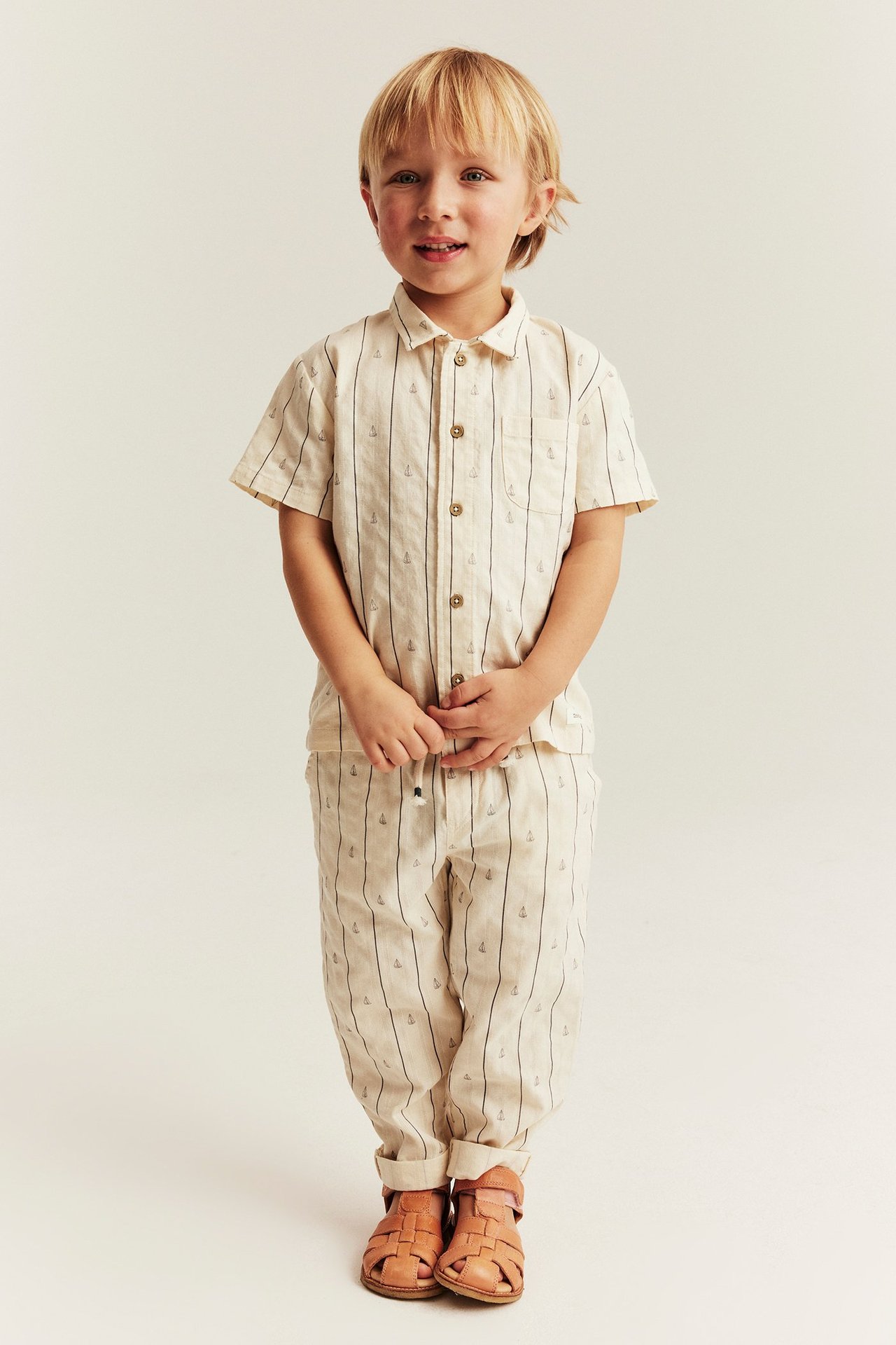 Vorderansicht: Kind in off-white gestreiften Baumwollhosen mit Segelbooten, passendem Hemd und Sandalen.