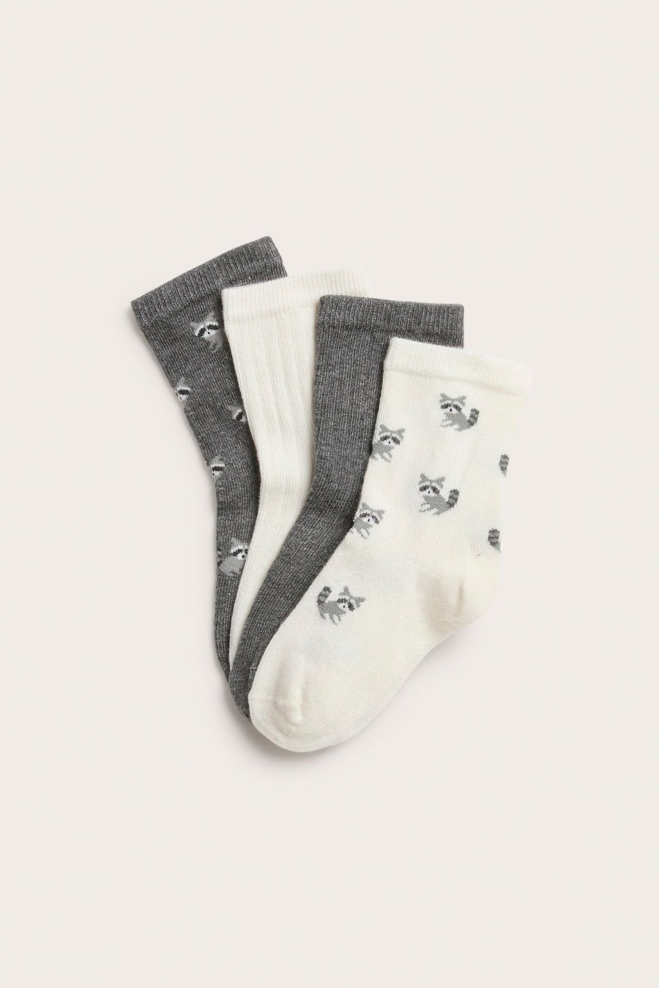 Vorderansicht von vier Kindersocken: zwei graue, zwei cremefarbene. Zwei mit Waschbär-Muster, zwei unifarben gerippt.