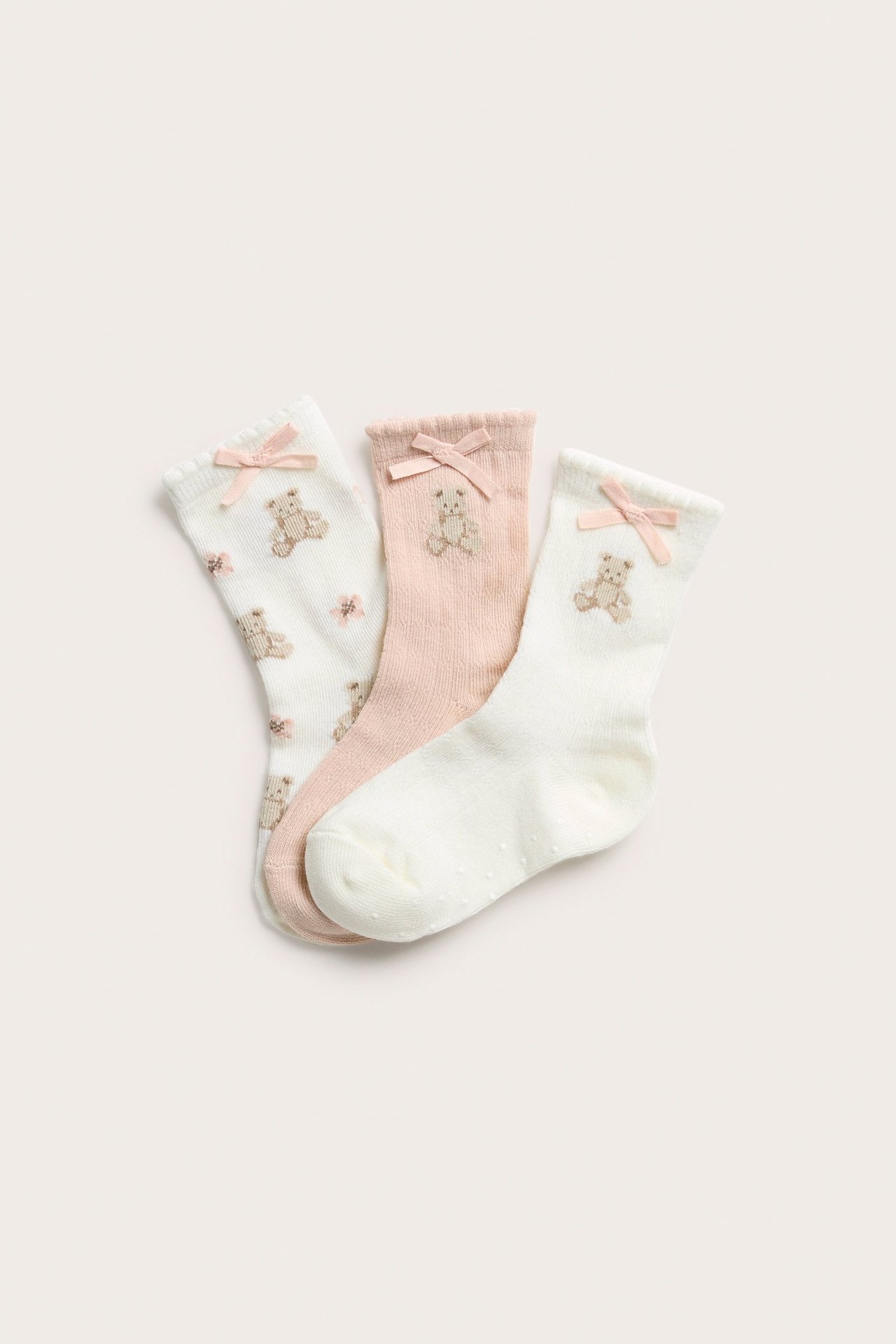 Draufsicht auf drei Paar cremeweiße und hellrosa Babysocken mit Teddybär-Stickerei und rosa Schleifen.