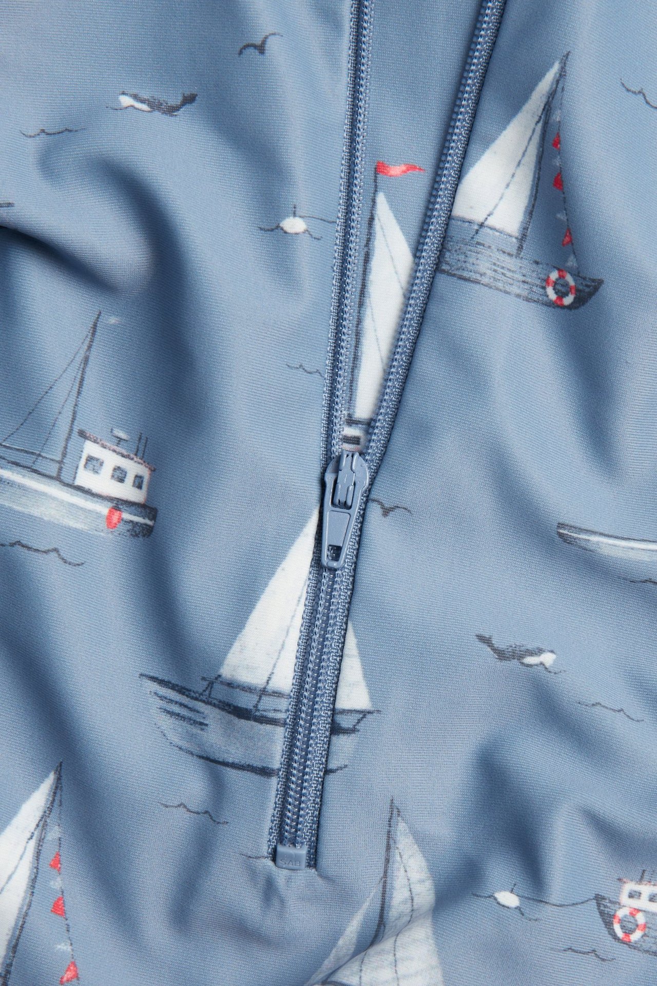 Nahaufnahme eines blauen Baby/Kinder UV-Rompers mit weißem Bootsmuster und sichtbarem blauen Reißverschluss.