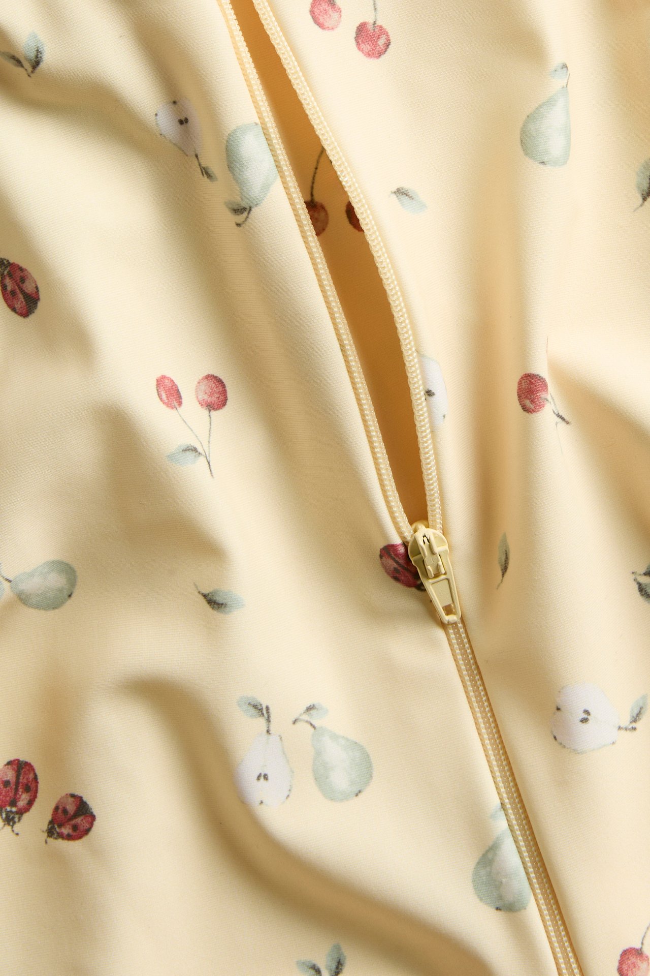 Close-up of a yellow patterned UV suit with cherries, pears, and ladybugs, featuring a zipper.