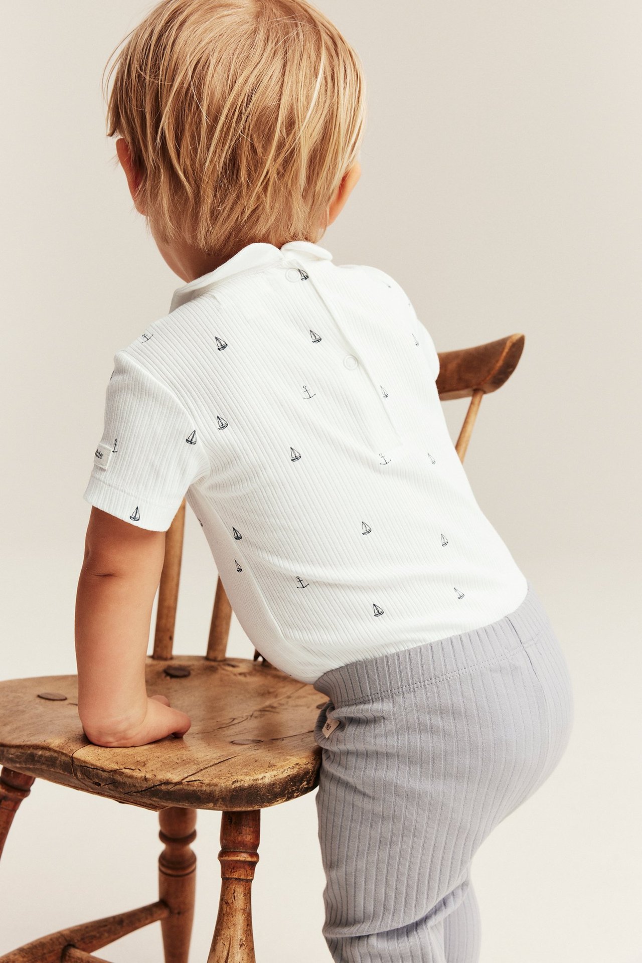 Rückenansicht eines Babys in einem cremefarbenen gerippten Kurzarmbody mit Segelbooten und grauen Ripp-Leggings.