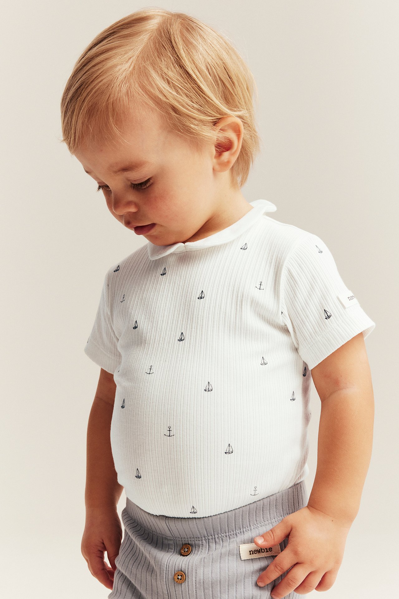 Front view of baby wearing off-white ribbed short-sleeve body with collar, marine pattern, grey pants.