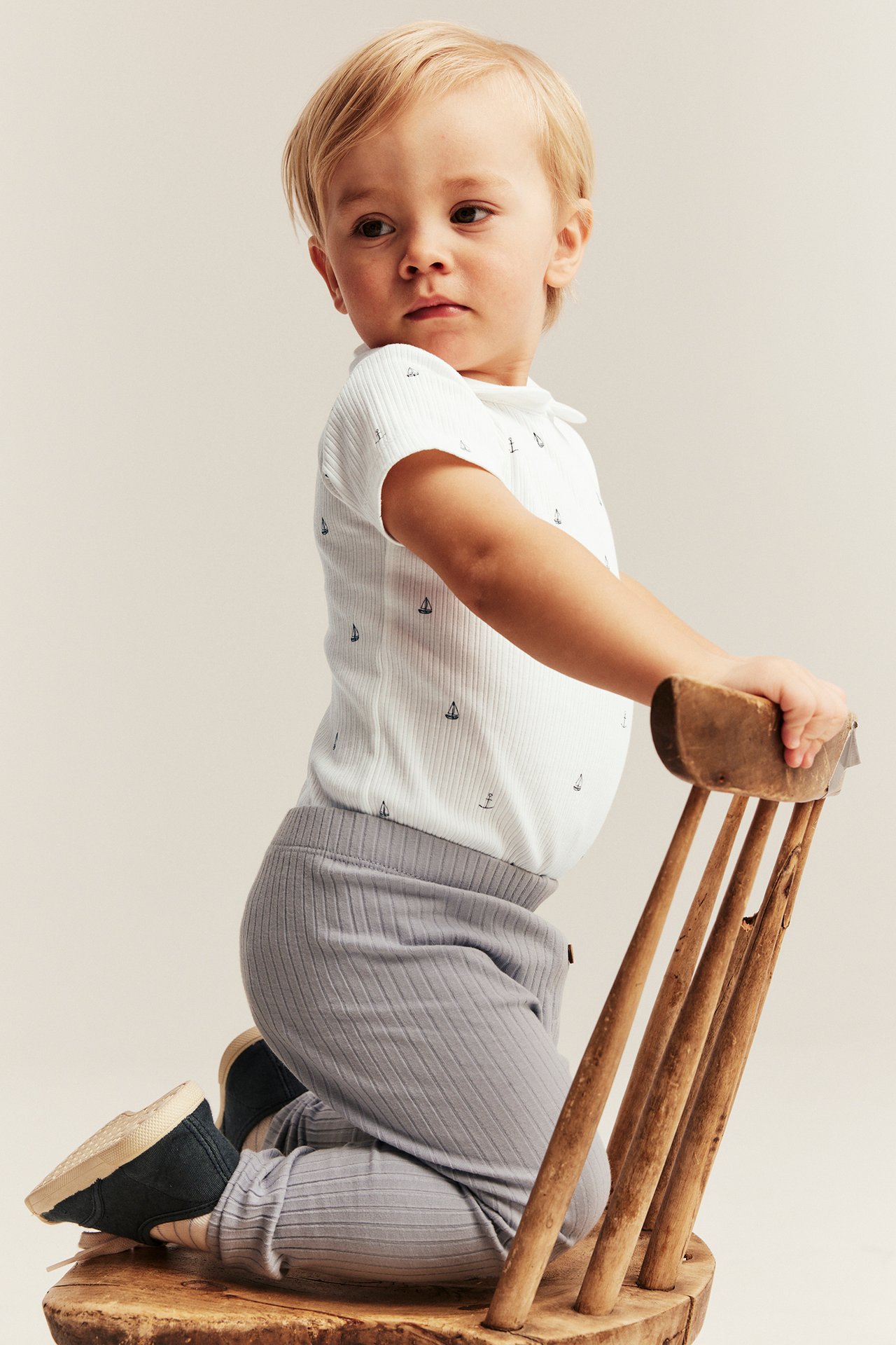 Seitenansicht eines Babys in einem cremeweißen gerippten Kurzarm-Body mit Segelbootmuster und grauen Leggings, kniend.