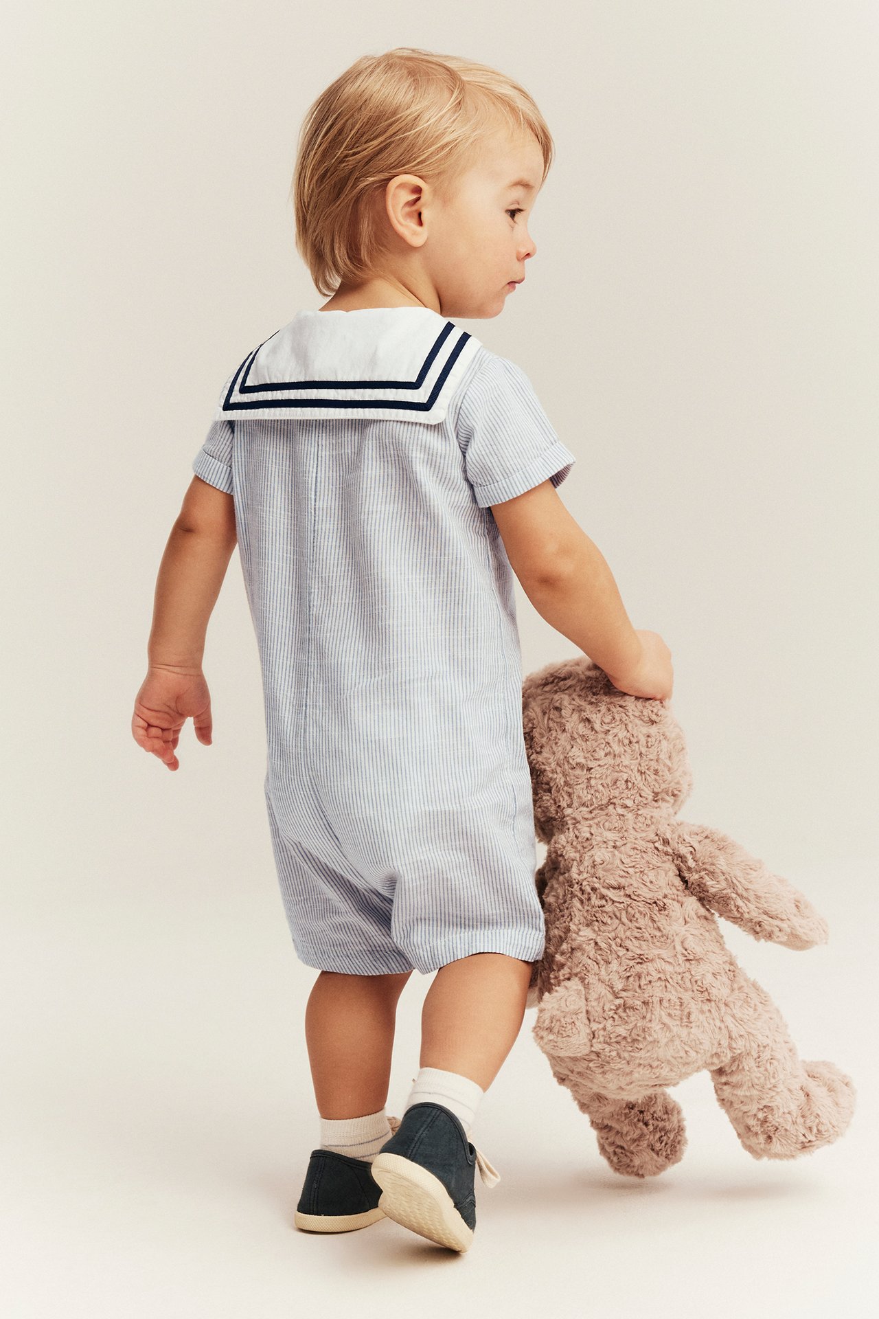 Back-side view: Baby in blue striped sailor romper, white socks, dark blue shoes, holding brown teddy bear.