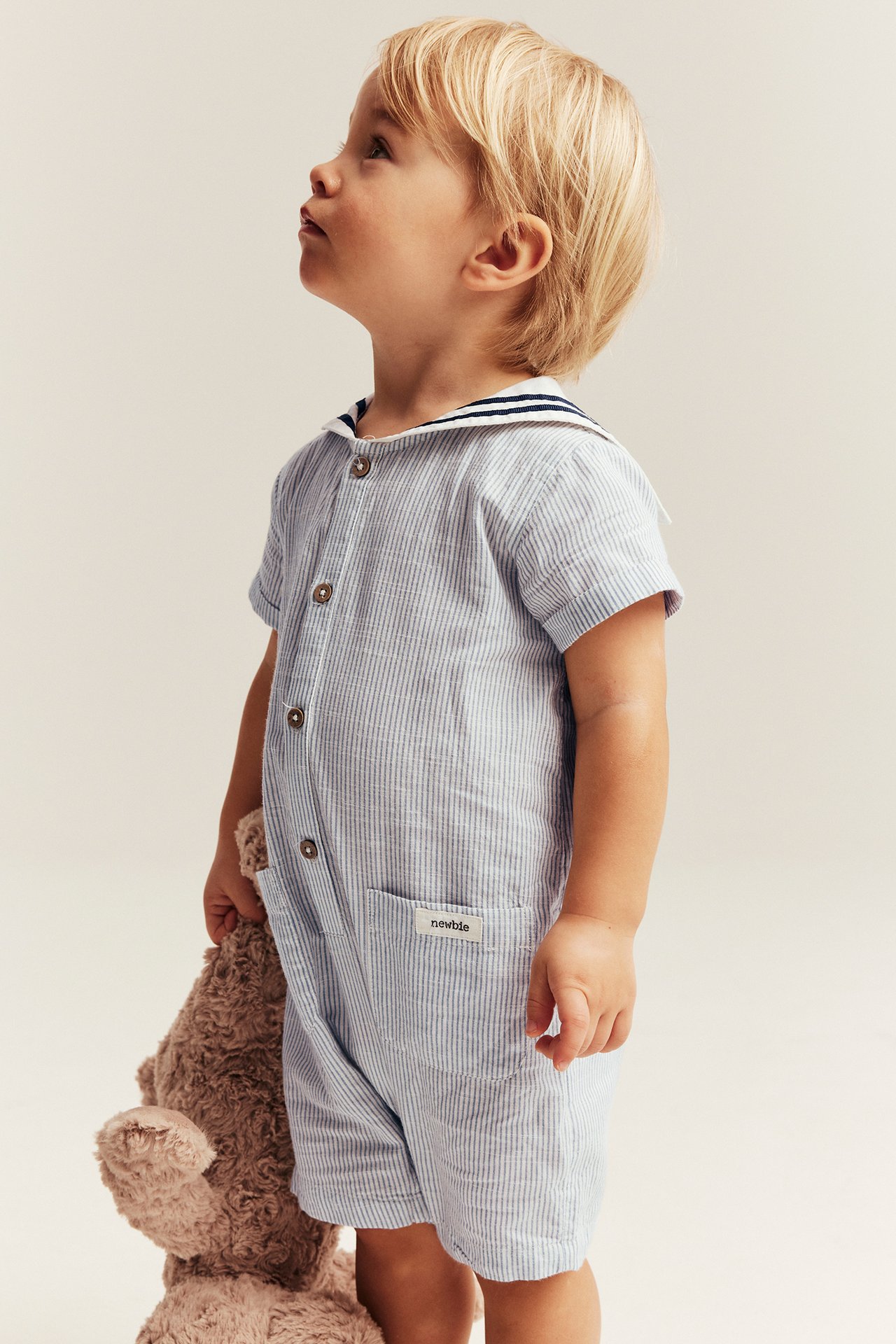 Side view: baby wears a light blue striped cotton romper with sailor collar, holding a brown teddy bear.
