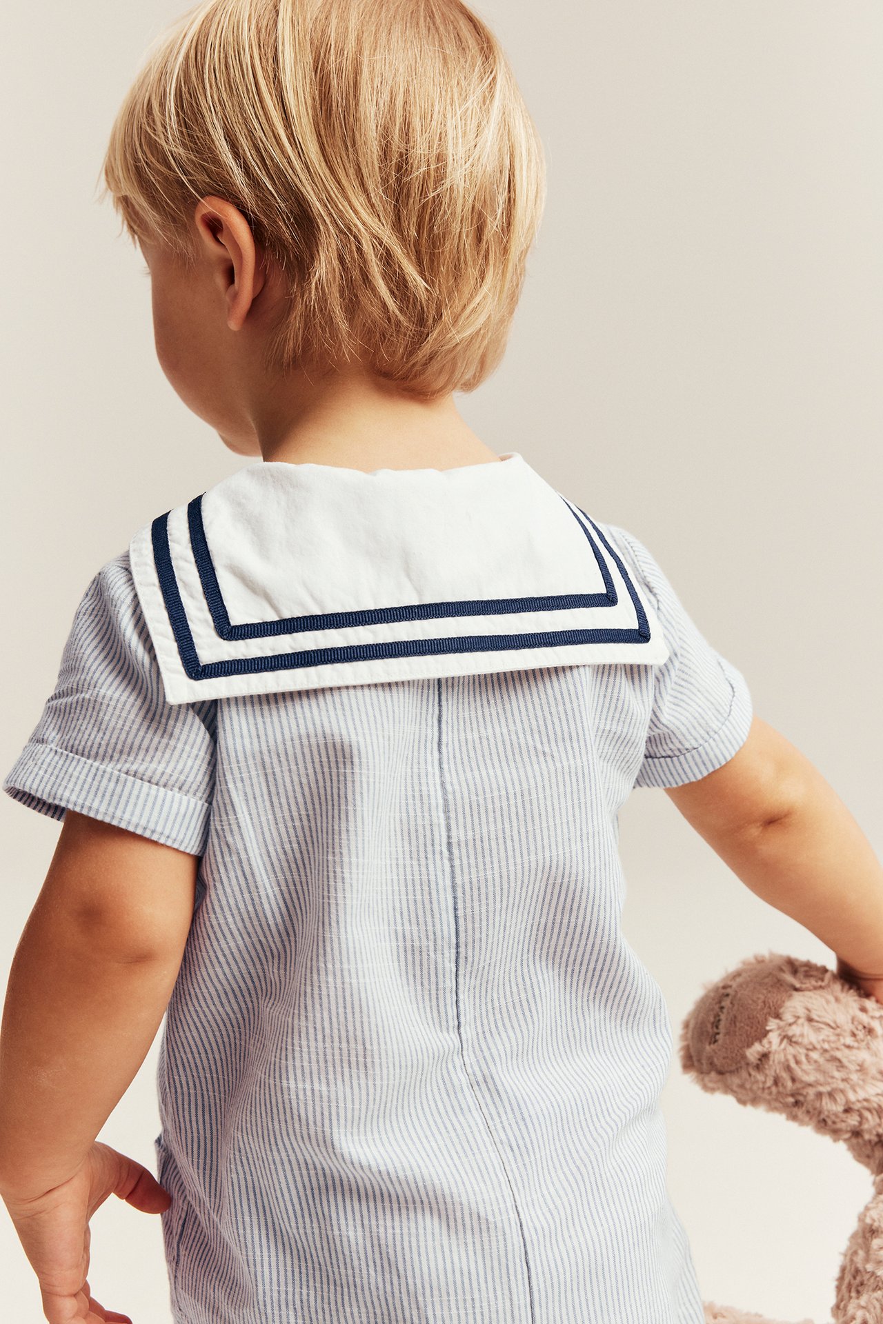 Back view of a baby wearing a blue striped woven romper with a white sailor collar, holding a plush toy.