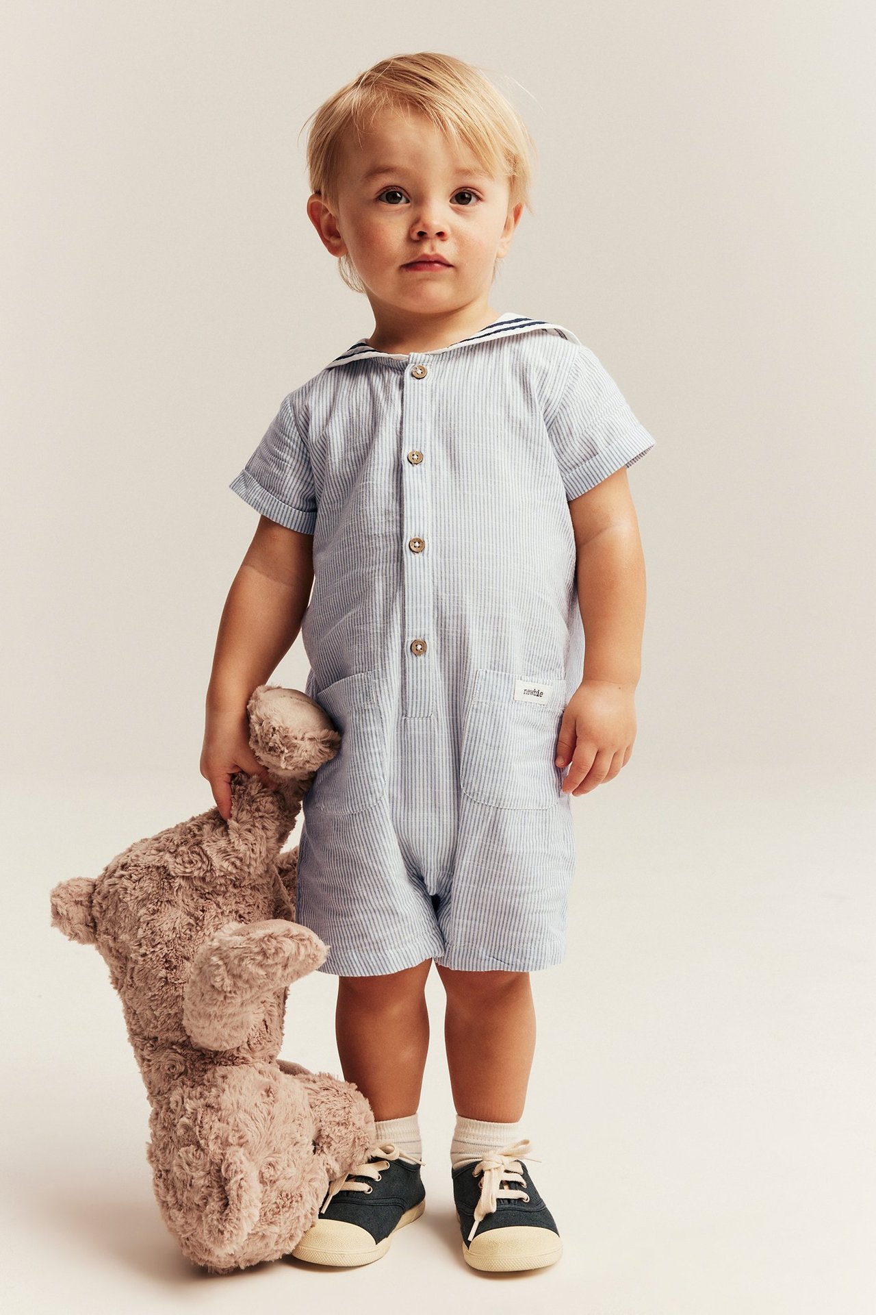 Front view: Baby in light blue striped romper, sailor collar. Holding brown teddy bear, dark blue sneakers.