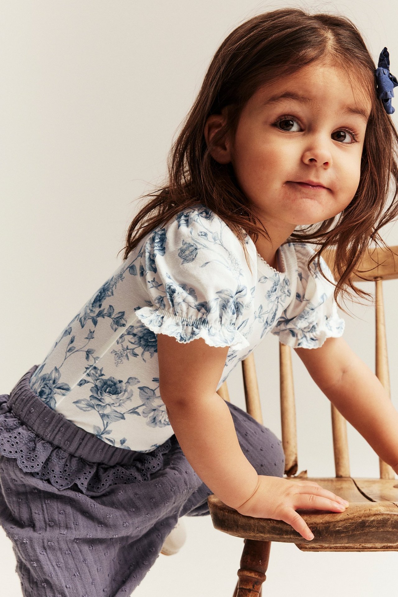 Vorderansicht eines Kindes in einem cremefarbenen geblümten Kurzarm-Body, dunklen Hosen und einer blauen Haarspange.