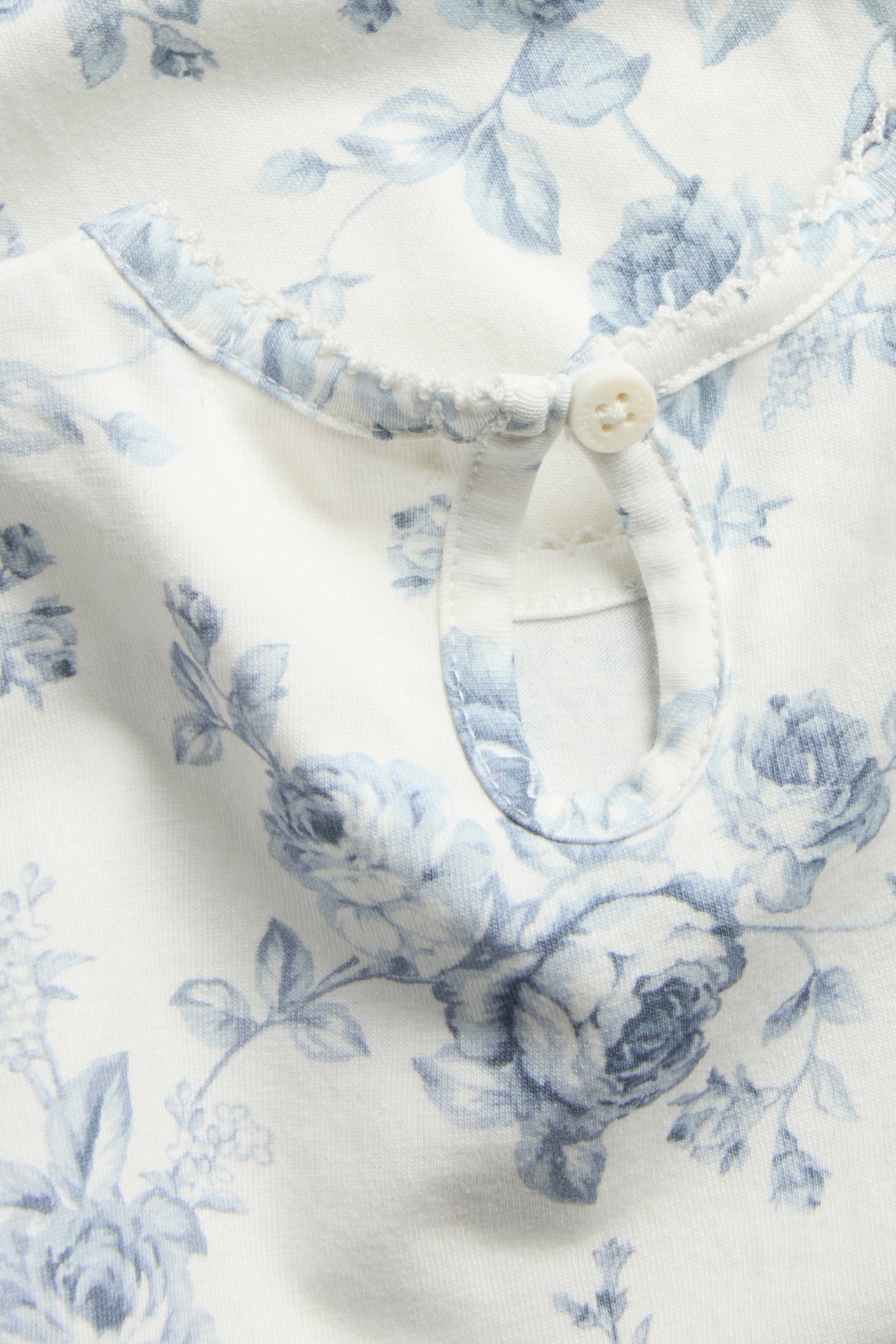 Nahaufnahme eines cremeweißen Baby-Bodys mit blauem Blumenmuster, Schlüssellochausschnitt und Knopf.