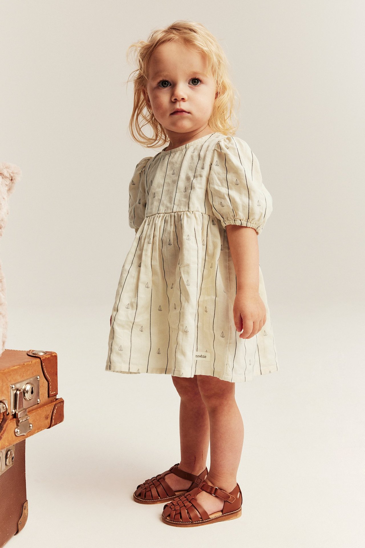 Vorderansicht: Baby-Mädchen in einem weißen gestreiften Baumwollkleid mit Segelbooten, Puffärmeln und braunen Sandalen.