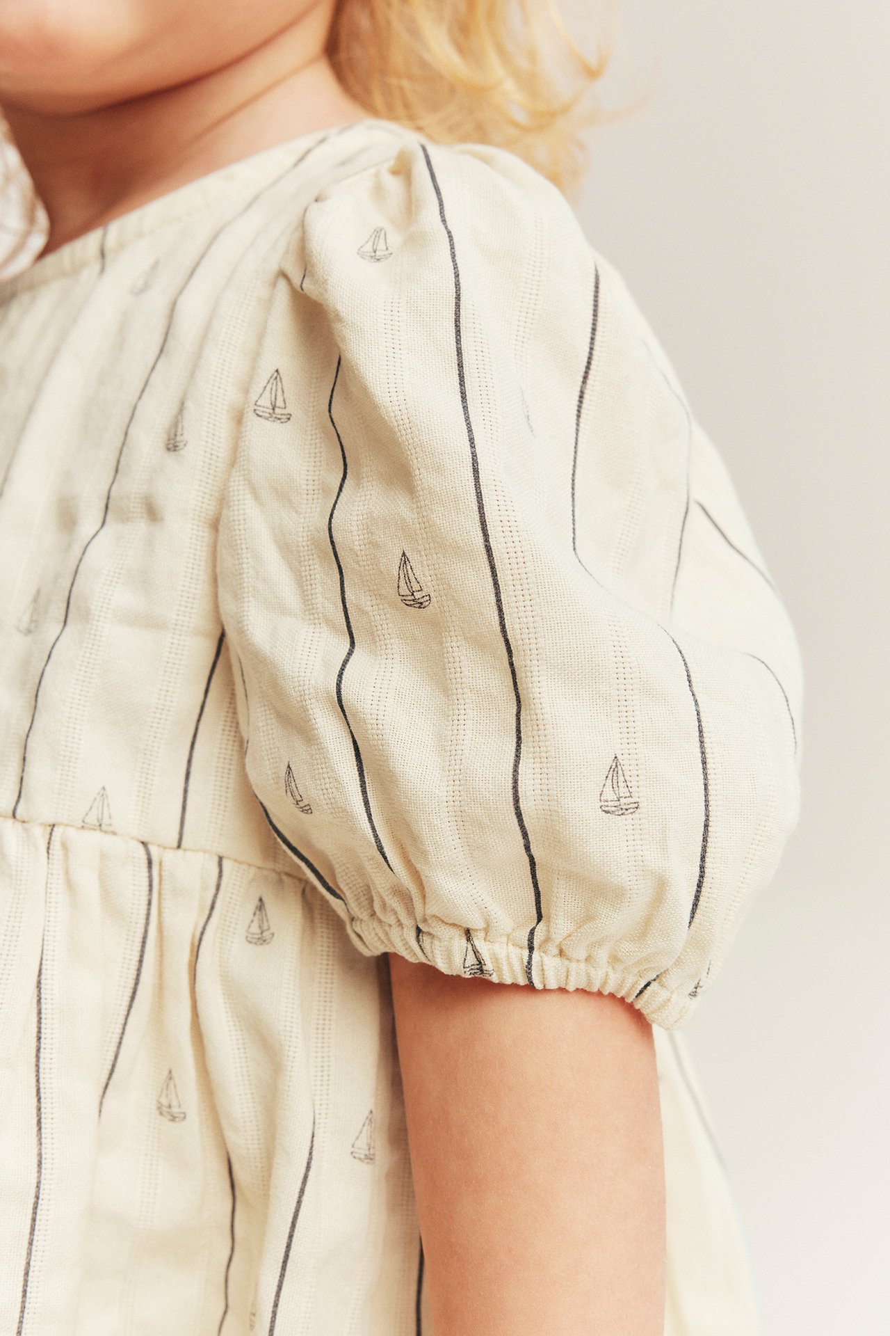 Close-up of a kid wearing a white cotton striped dress with printed sailboats and elasticized puff sleeves.