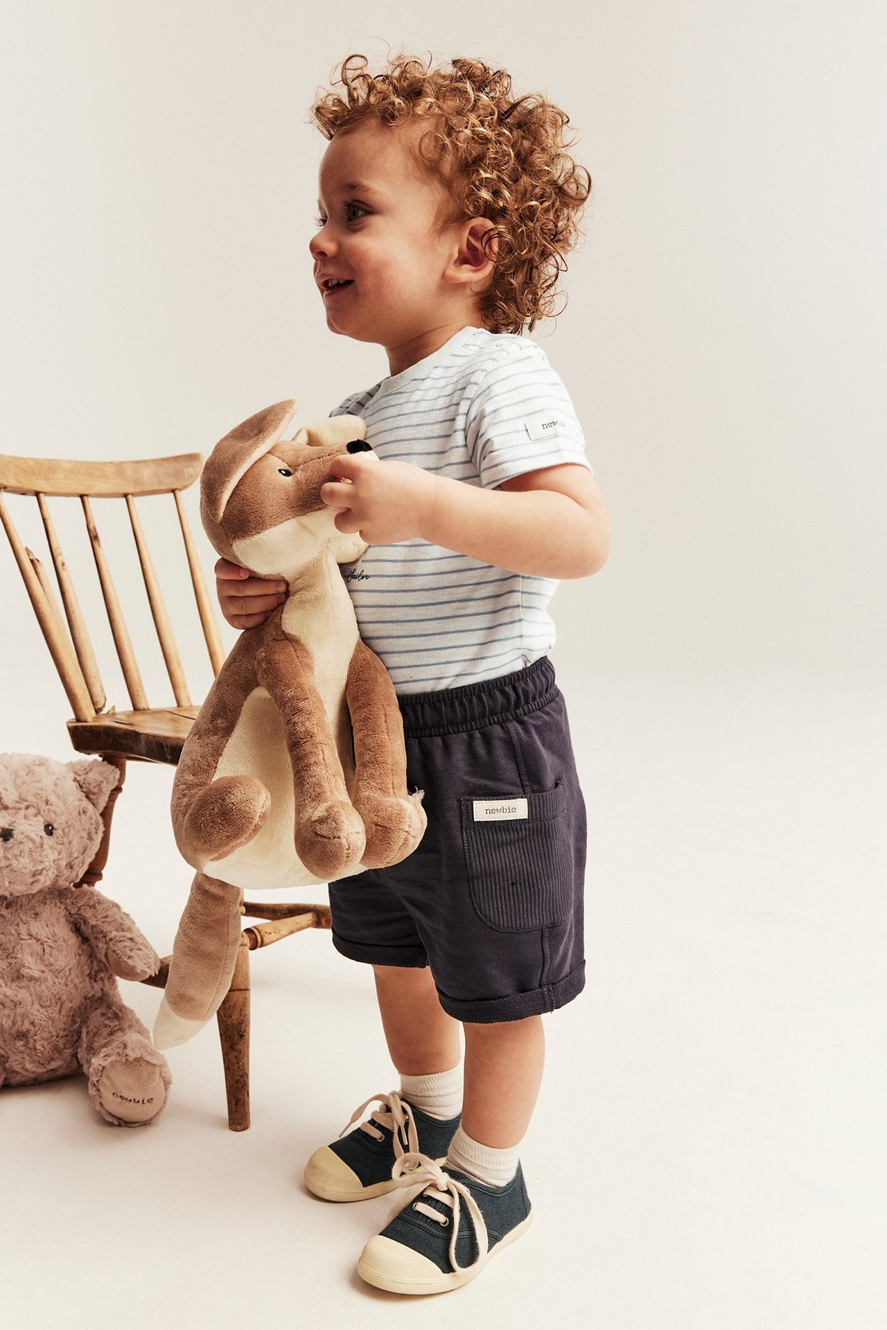 Side view of a kid in an off-white striped body with teddy application, dark blue shorts, and shoes.