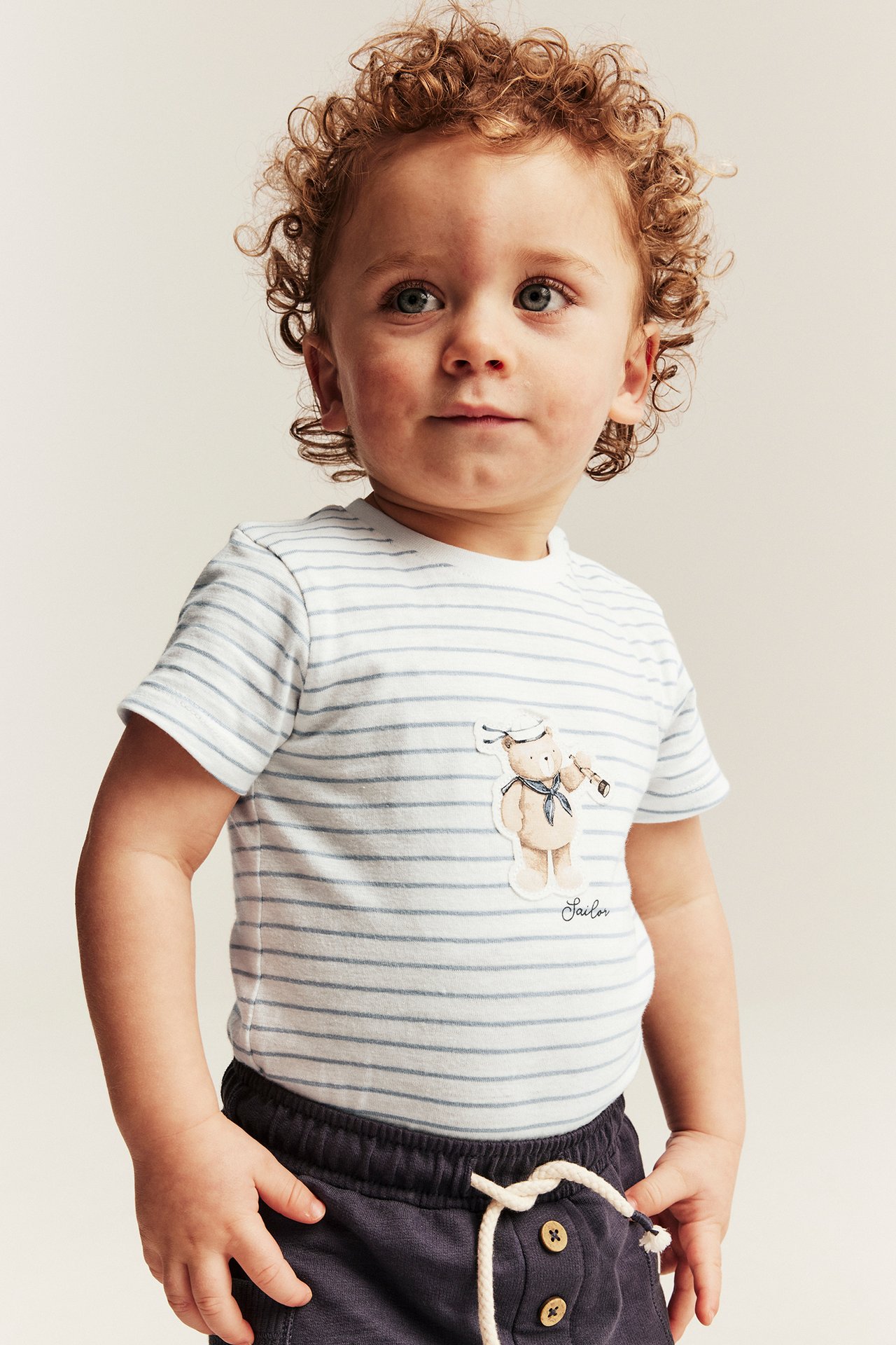 Front view of a baby wearing an offwhite striped short-sleeved cotton bodysuit with a teddy bear appliqué and dark shorts.