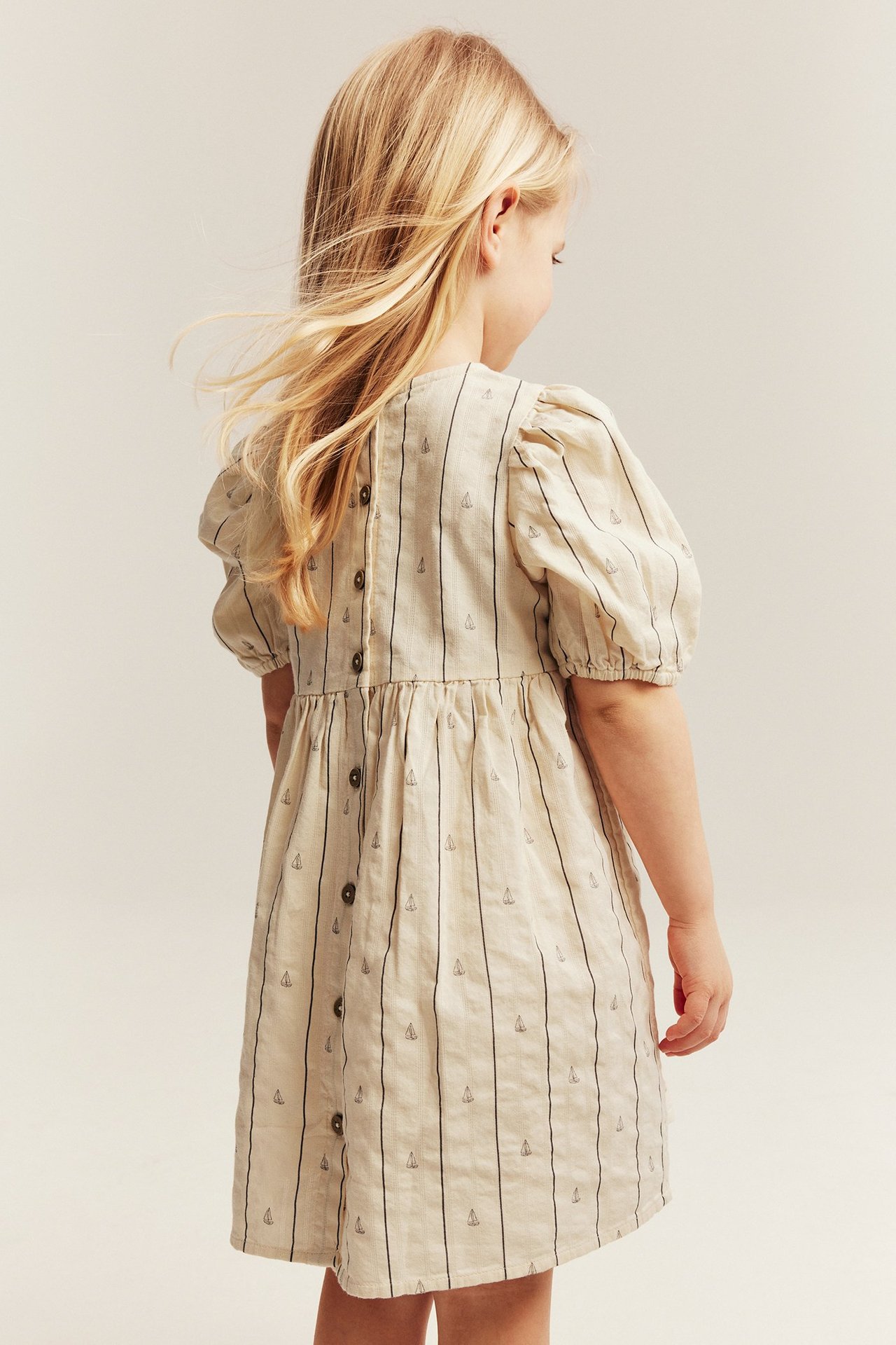 Back view of a kid wearing a white cotton dress with vertical stripes, sailboats, puff sleeves, and back buttons.