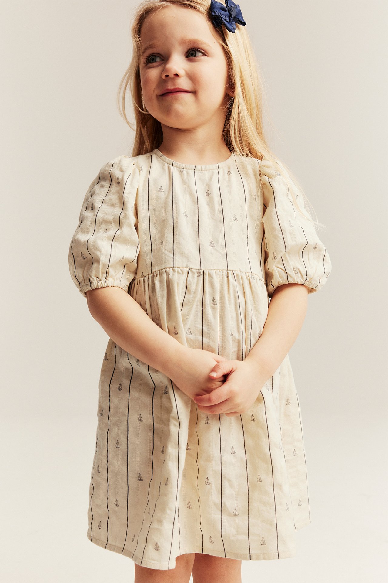 Front view: Kid in off-white striped cotton dress, boat print, puff sleeves, and a navy bow clip.