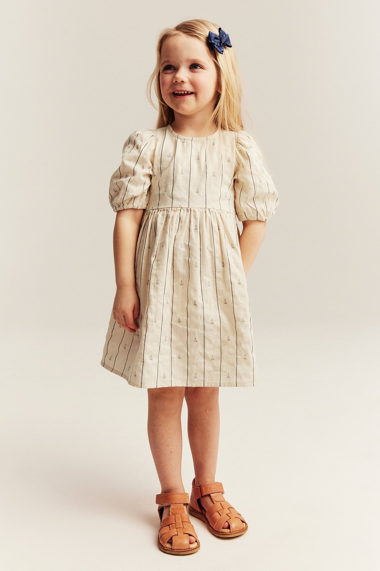 Front view: Kid girl in white striped boat print dress, dark blue bow, and brown sandals.