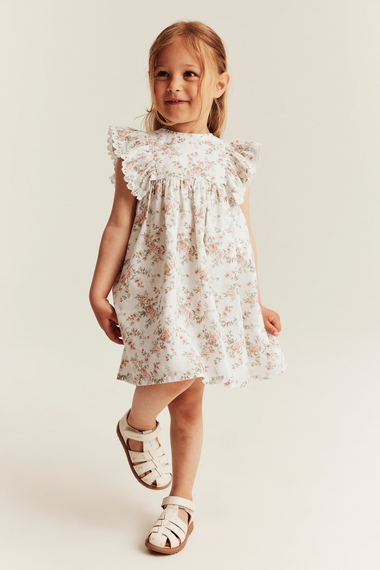 Vorderansicht eines Kindes in einem cremeweißen geblümten Rüschenkleid mit Spitzenkanten und cremeweißen Sandalen.