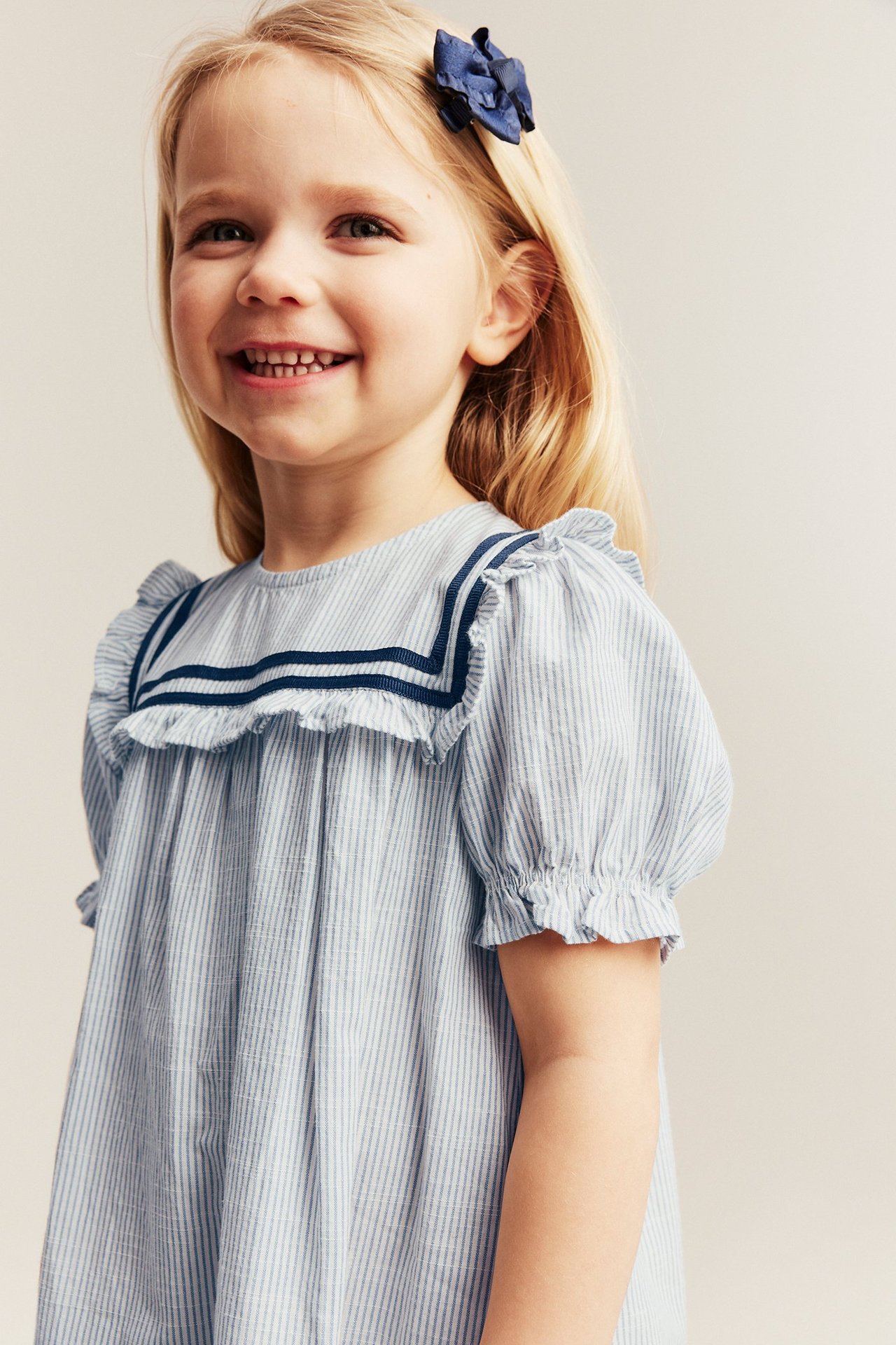 Front view, kid in blue striped cotton sailor dress with puff sleeves, wearing a blue bow clip.