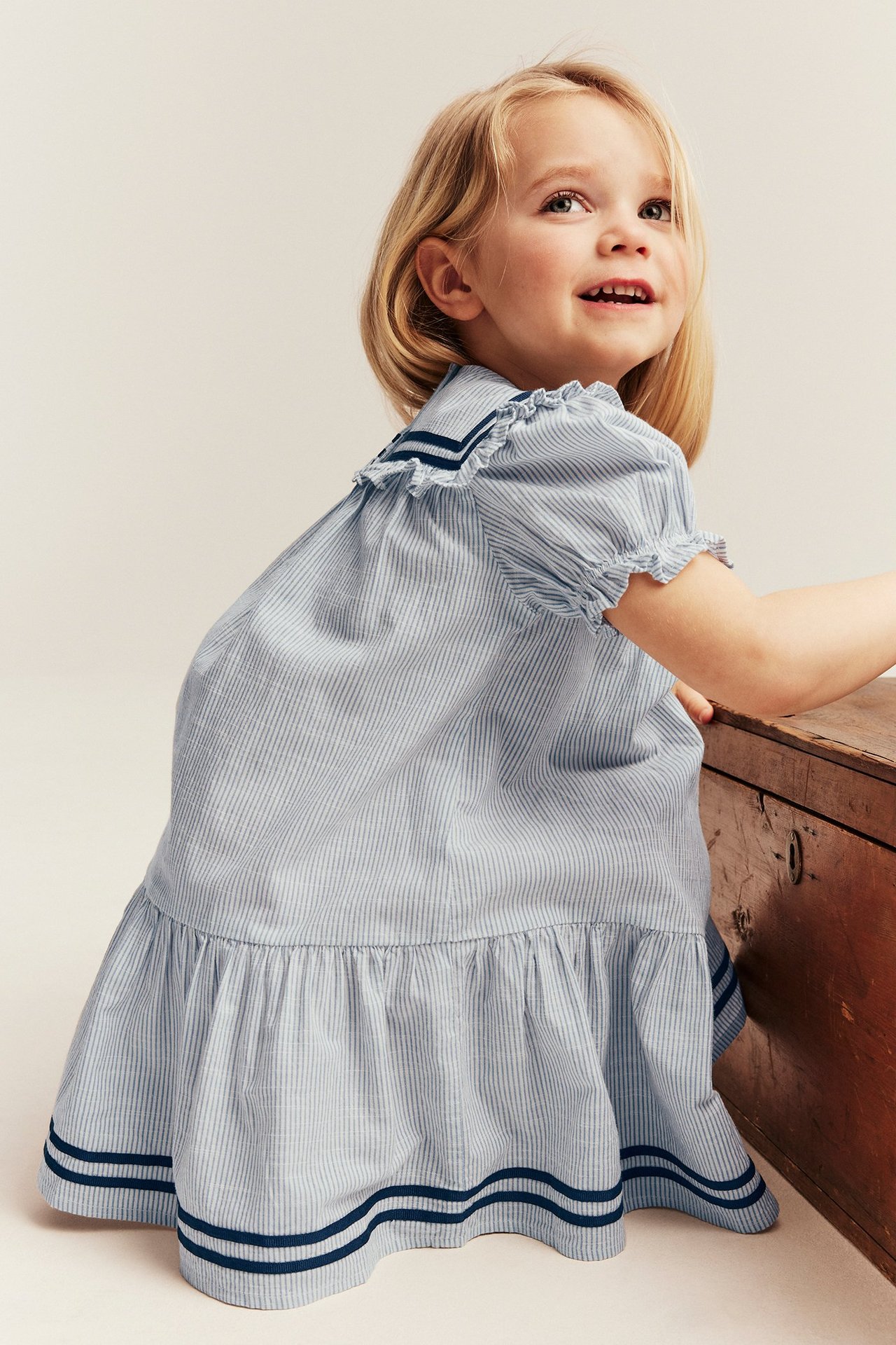 Side view of a kid wearing a light blue striped cotton sailor dress with puff sleeves and blue bands.