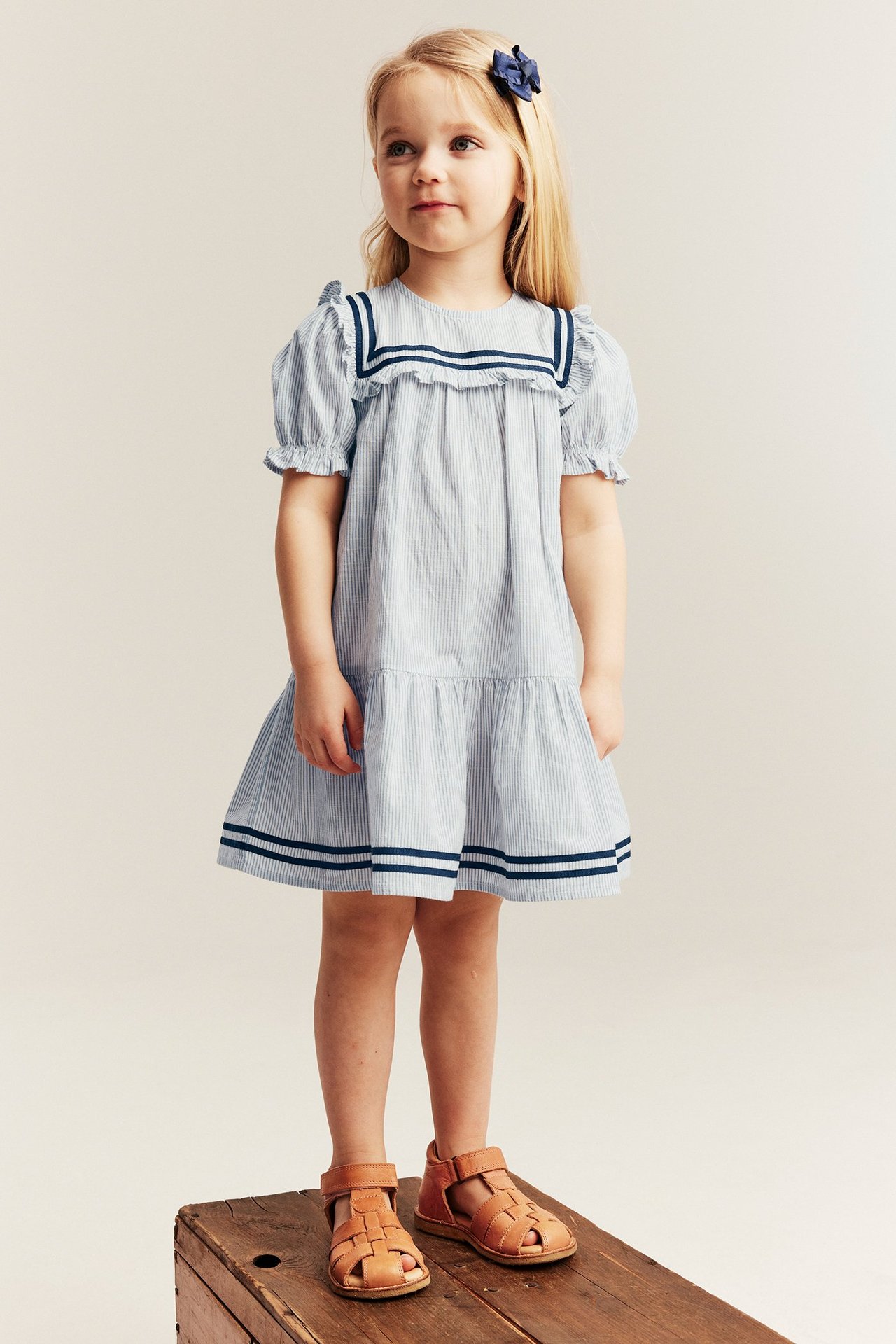 Kid in light blue striped cotton sailor dress with puff sleeves and brown sandals, front view.