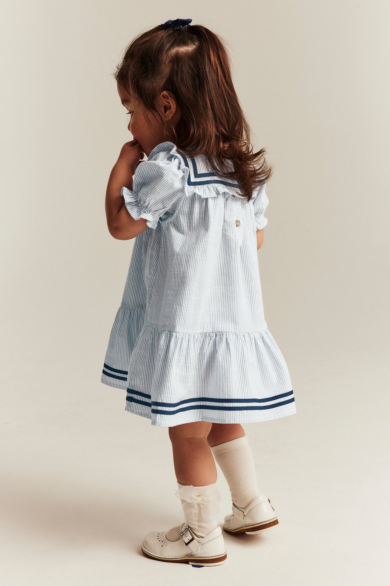 Back view of a kid in a light blue striped sailor dress with puff sleeves, white socks, and white shoes.